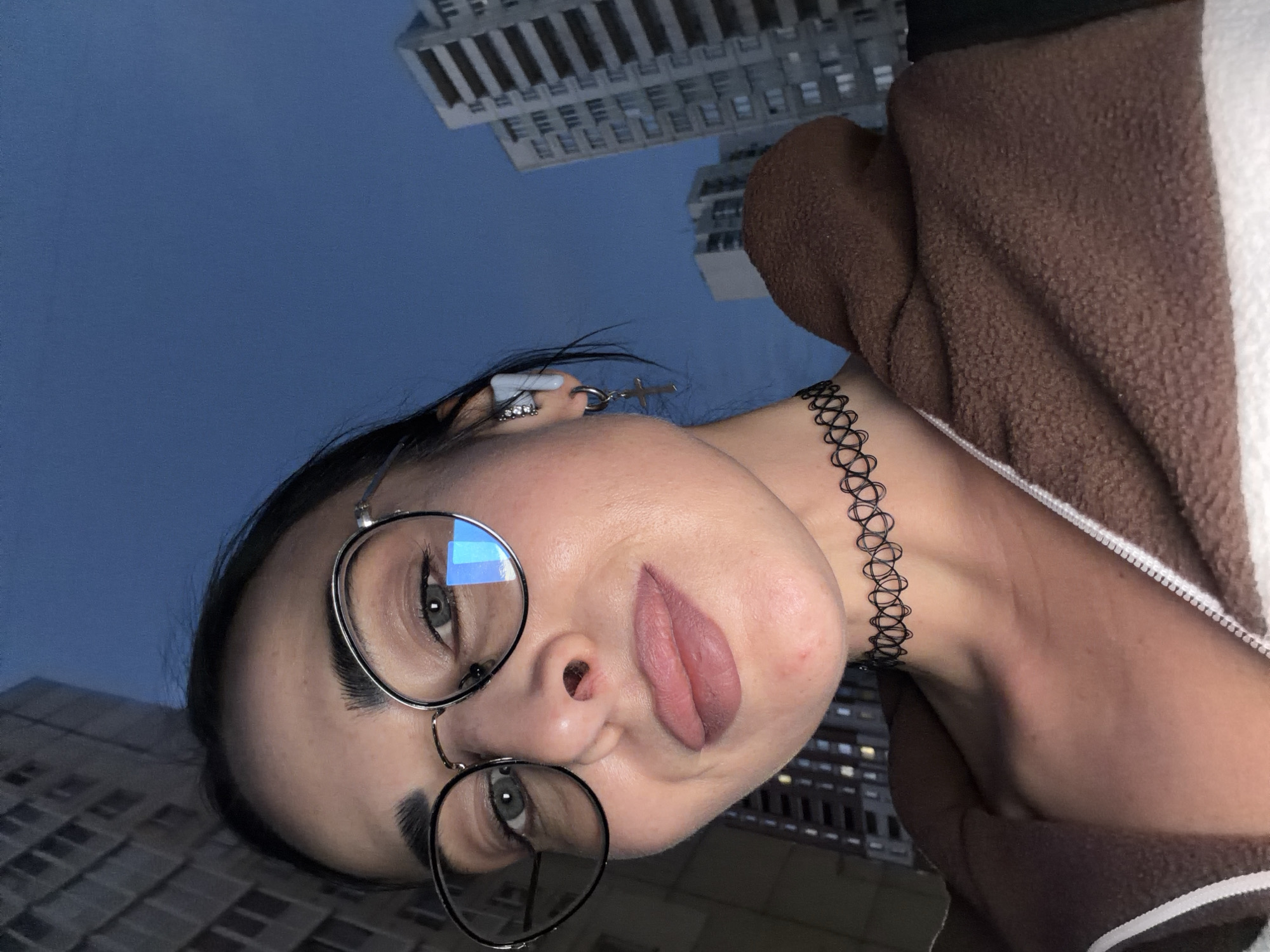 A low-angle, diagonal portrait features a young adult woman gazing upwards towards the camera, set against a backdrop of a deep blue sky and urban high-rise buildings. The woman, visible from the upper chest, has dark hair, dark eyebrows, and light-colored eyes. She wears round, metallic-rimmed glasses that reflect a blue light. Her lips are a muted pink, slightly parted, suggesting permanent lip blush or bold lipstick.
She is accessorized with a black, tattoo-style choker necklace. Her right ear displays multiple piercings, including a small silver cross dangling from one and a small white, cylindrical object, possibly an earpiece or an intricate earring, from another. She is clad in a textured, brown hooded jacket with a visible white lining or trim.
The background shows several tall, modern buildings, with some windows dimly lit, rising into the dark blue sky. The sky's hue and the artificial light from the structures indicate the photograph was taken during the evening or night. The overall scene conveys a casual, urban portrait. The location context provided is Saint Petersburg, Russia. No visible text is present in the image.