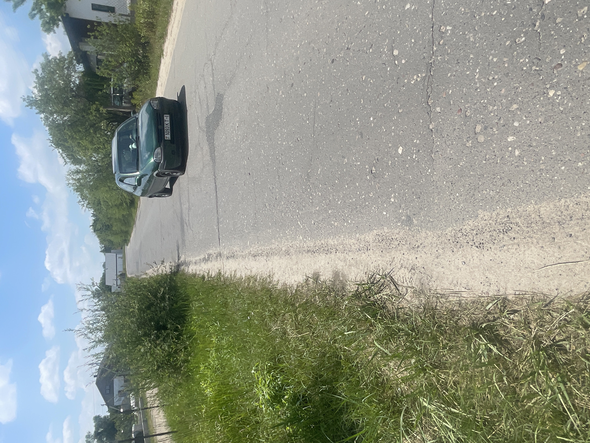 A green Opel Corsa is driving down a two-lane asphalt road in a rural area, possibly in Мухавецкі сельскі Савет, Belarus, given the license plate format. The car has a license plate that reads "1093 KT-1". The road is lined on the left by a grassy verge and then more grass and bushes, with houses and trees visible in the background. The sky is blue with scattered white clouds. The day is bright and sunny.