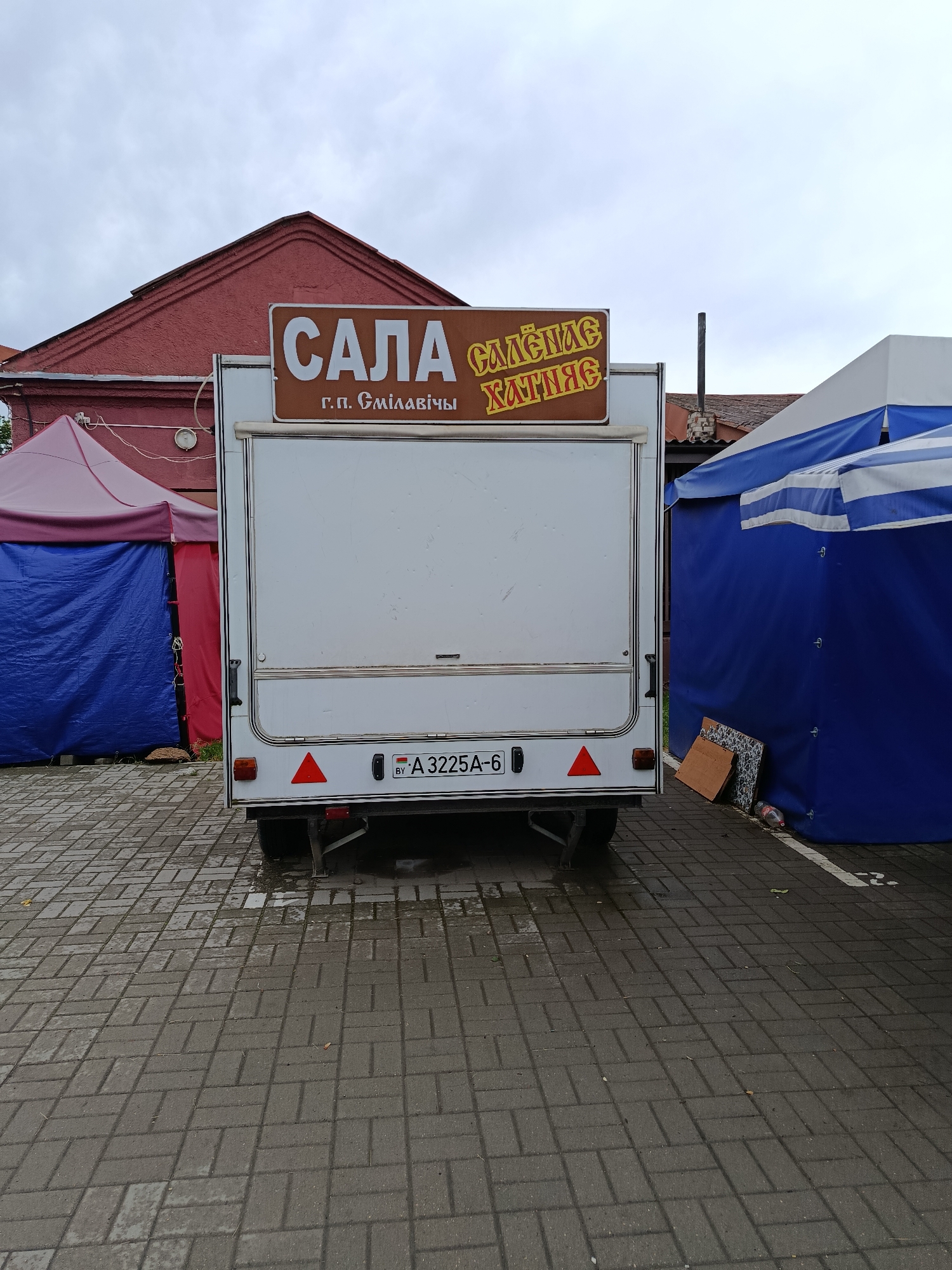 The image depicts a white food trailer parked on a wet, paved surface in what appears to be an outdoor market setting. A red brick building is visible in the background to the left, with a red and pink tent set up in front of it. To the right of the trailer, a blue and white striped tent is visible.
The trailer has a sign at the top which reads "САЛА" in large white letters. Below this, in smaller text, it says "г.п. Смілавічы", indicating the location as Smilavichy rural council in Belarus. To the right of "САЛА", a yellow sign with red lettering reads "СЛАДКАЯ ХАТКА".
A license plate on the back of the trailer reads "BY A 3225A-6". There are two red triangular reflectors on the back of the trailer. A black handle is visible on the side of the trailer's rear door.
The ground is paved with grey interlocking bricks and appears wet, suggesting recent rain. A discarded plastic bottle and some miscellaneous items are visible near the blue tent on the right. The sky is overcast. There are no people visible in the image.