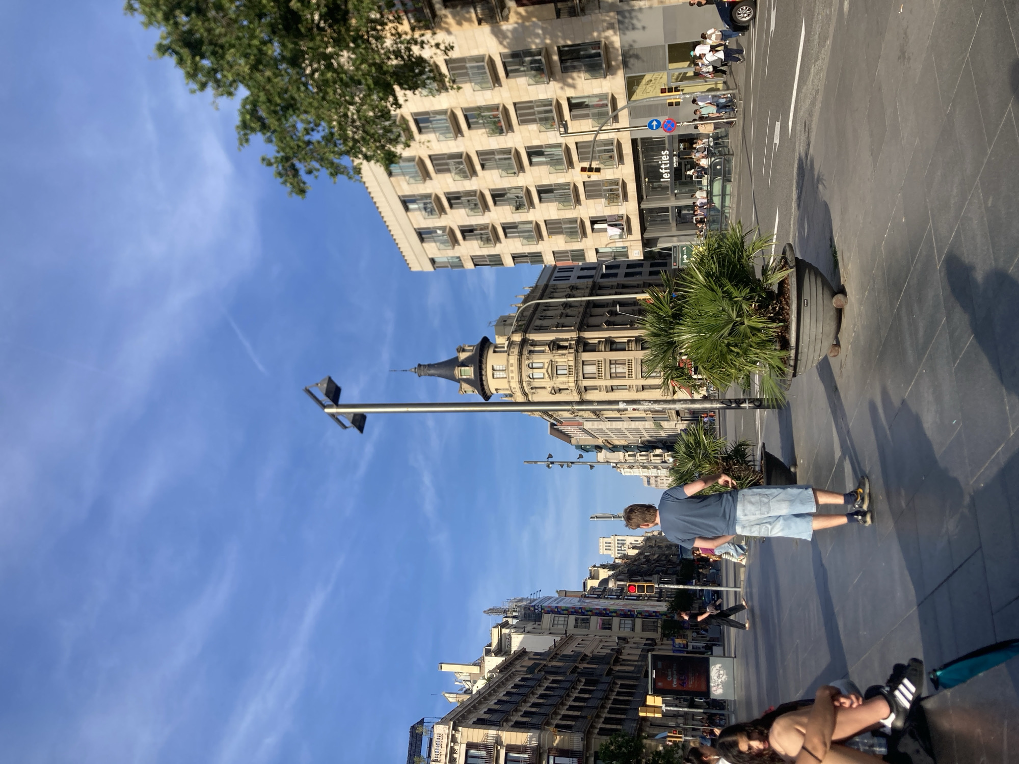 The image shows a street scene in Barcelona, Spain. A man in a blue t-shirt and jean shorts is walking on the sidewalk. There is a large potted plant next to the sidewalk. Buildings line both sides of the street, and there are traffic lights and street signs. There are other people walking on the street and sidewalk. The sky is blue with some clouds. A store called "lefties" is visible in the background.