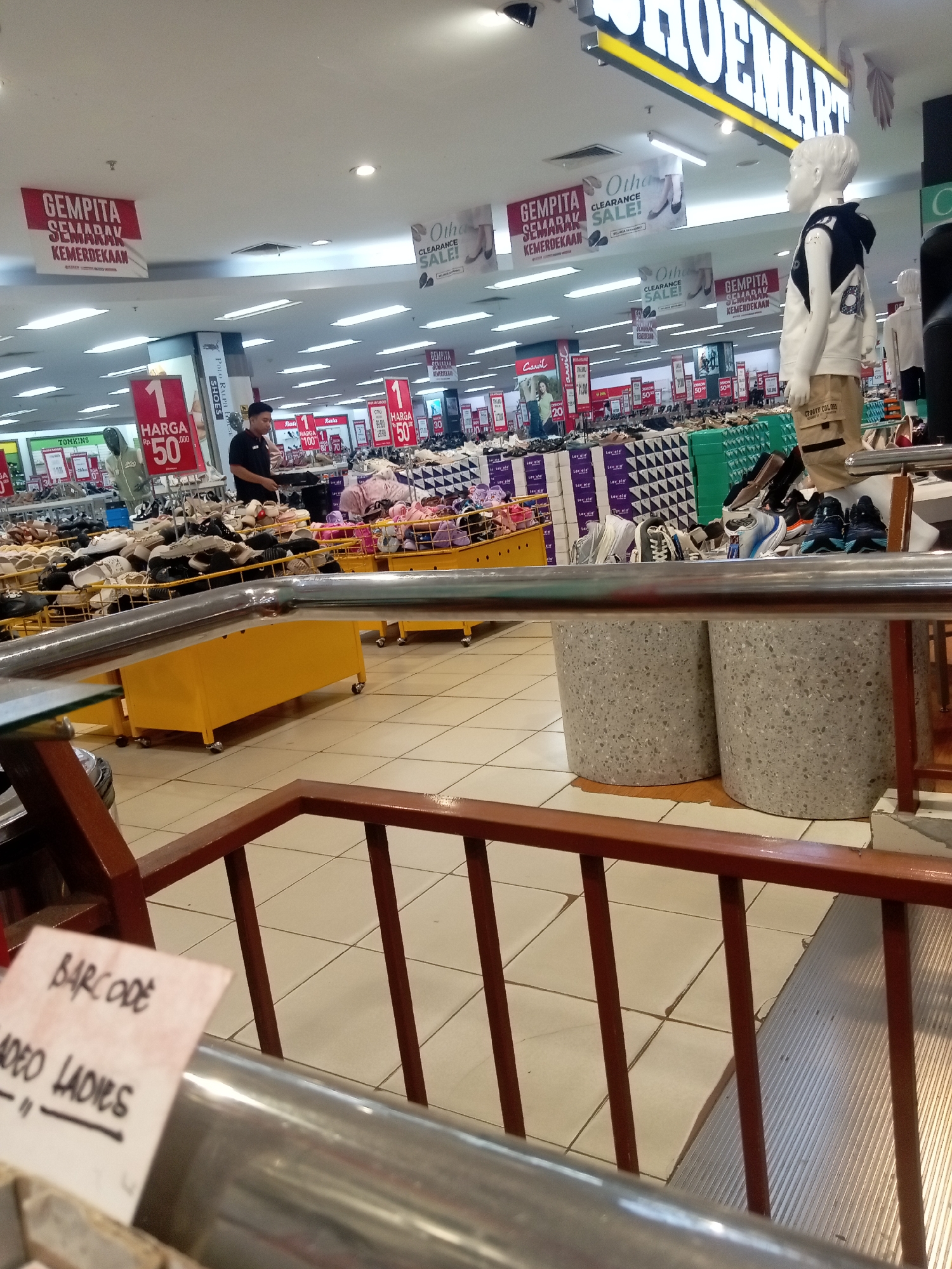 The image depicts the interior of a shoe store, likely Shoemart, located in Batam, Indonesia, evidenced by prominent signage. The store features a wide selection of shoes displayed on shelves and in baskets. Several red banners with white text are visible, announcing "GEMPITA SEMARAK KEMERDEKAAN" and "CLEARANCE SALE!". A male store employee in a black shirt is standing near a display of shoes. Mannequins dressed in casual clothing are also present. In the foreground, a metal railing and wooden structure are visible, suggesting a mezzanine or elevated viewing area. The floor is tiled, and the ceiling is illuminated by fluorescent lights. The overall atmosphere is that of a busy retail environment.