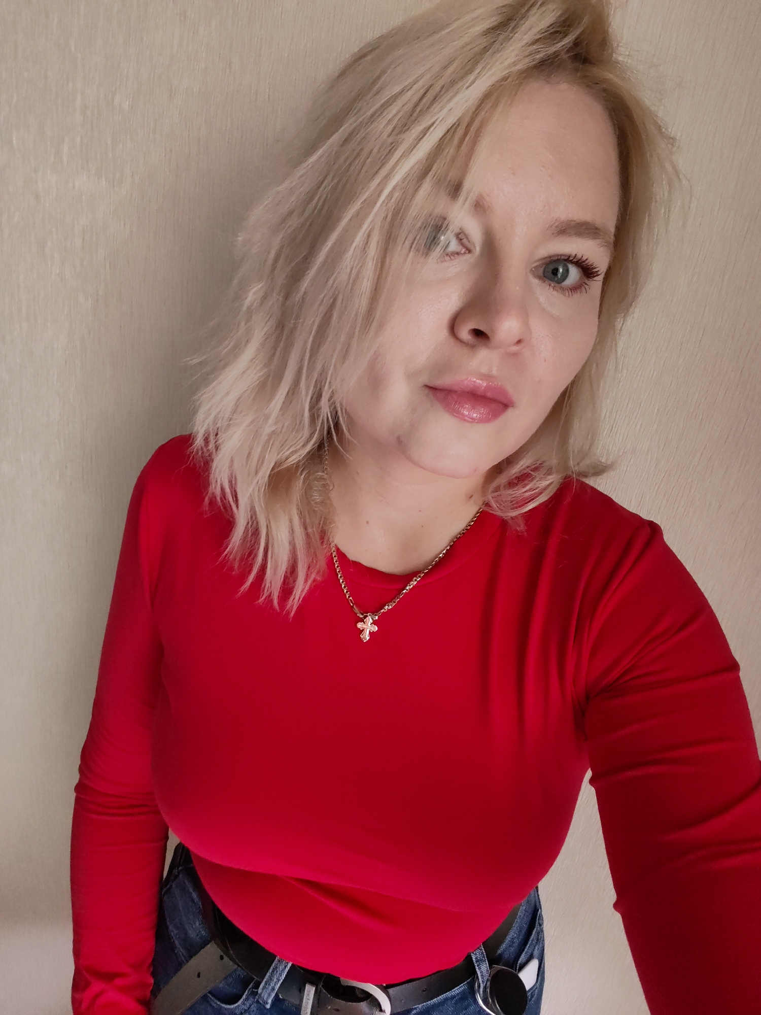 A blonde woman with blue eyes and fair skin is looking at the camera. She is wearing a red long-sleeved shirt and blue jeans. She has on a silver necklace with a small cross pendant. The background is a plain, textured wall. The image appears to be a selfie taken indoors, with no specific geographic markers to indicate a location in Izhevsk, Russia.