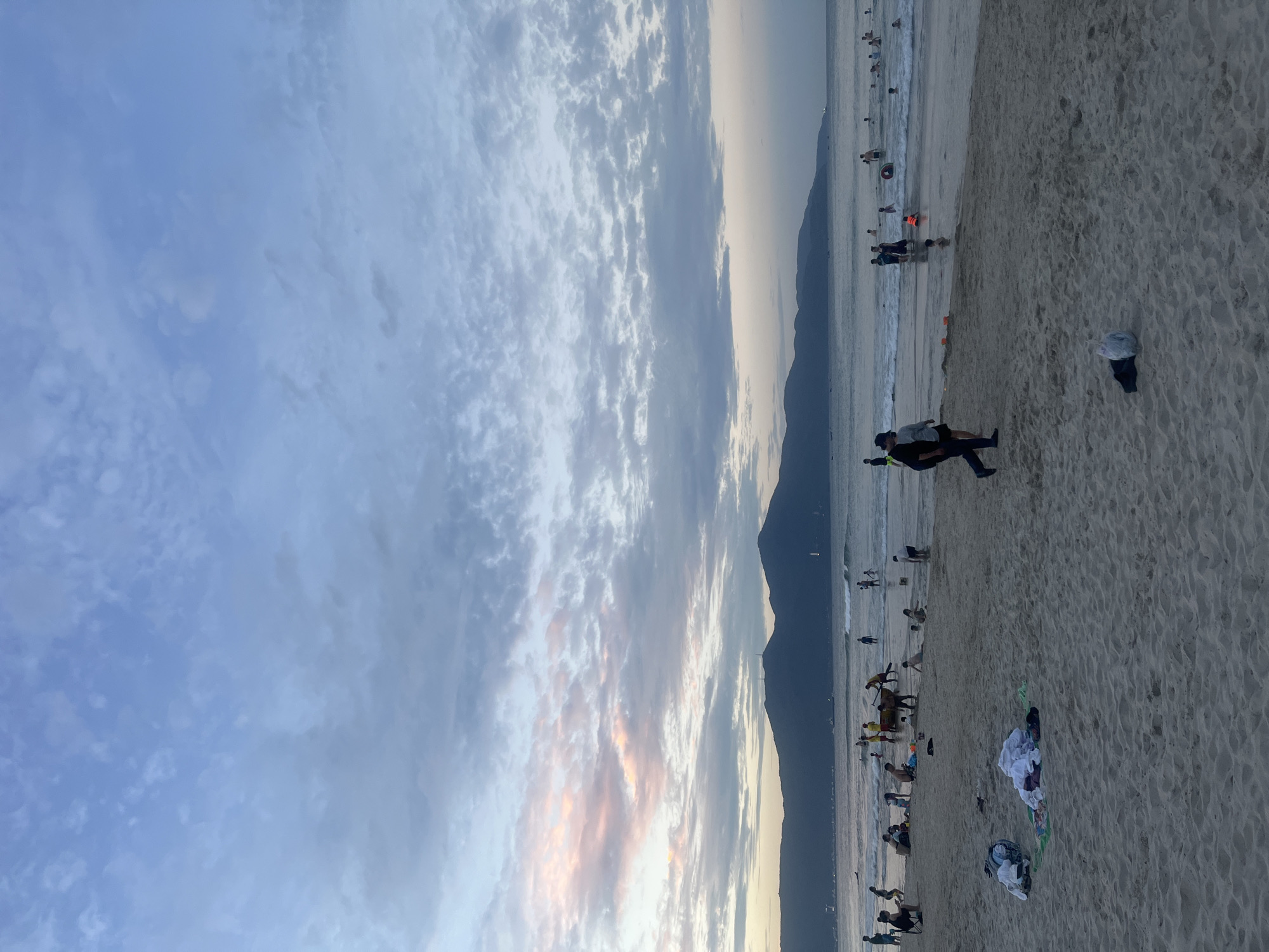 This is a photograph of a beach in Da Nang, Vietnam, likely taken in the late afternoon or early evening, given the soft light and the colors in the sky. The image is dominated by a wide expanse of sandy beach in the foreground, with several piles of clothing and bags scattered on the sand. In the middle ground, a calm sea stretches out, with numerous people wading, swimming, and playing in the shallow water. Some individuals are further out in the water, and a few appear to be on paddleboards or similar watercraft.
In the background, a range of dark, silhouetted mountains forms the horizon line, with a faint glow of sunset or twilight peeking through the clouds above them. The sky is a tapestry of blues and grays, with scattered clouds, some of which are tinged with pink and orange, suggesting the sun is low in the sky. The overall atmosphere is one of leisure and recreation, with a sense of community as people enjoy the beach.