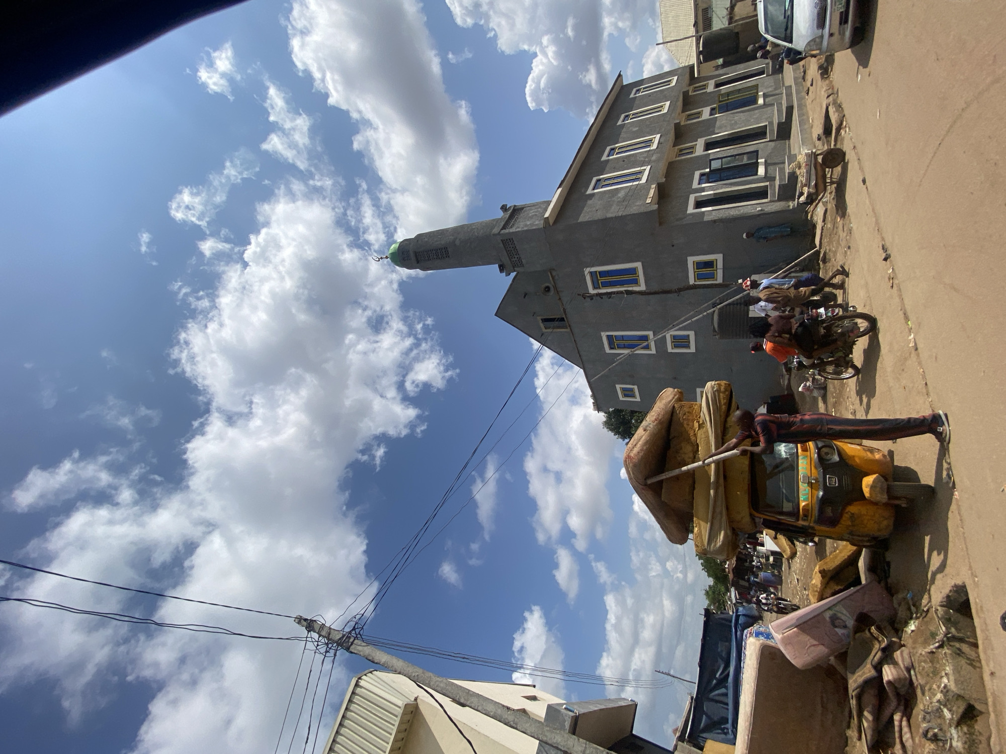 The image depicts a street scene in Ado, Nigeria. In the foreground, a yellow auto-rickshaw is heavily laden with mattresses, balanced precariously on top and secured with a pole. A man in a striped shirt and pants stands beside the auto-rickshaw, holding the pole. To the left, more mattresses are piled on the ground and leaning against a building.
Dominating the mid-ground is a gray mosque with a green dome and a minaret. The mosque has several rectangular windows with blue frames. Several people are visible near the mosque, including individuals walking and a group on a motorcycle. A silver car is parked on the right side of the road.
In the background, various buildings, including what appears to be another structure with a light-colored facade on the left, and a blue tarp are visible. The sky is blue with scattered white clouds, and power lines stretch across the frame. The overall environment suggests a bustling, possibly developing, urban or semi-urban area.