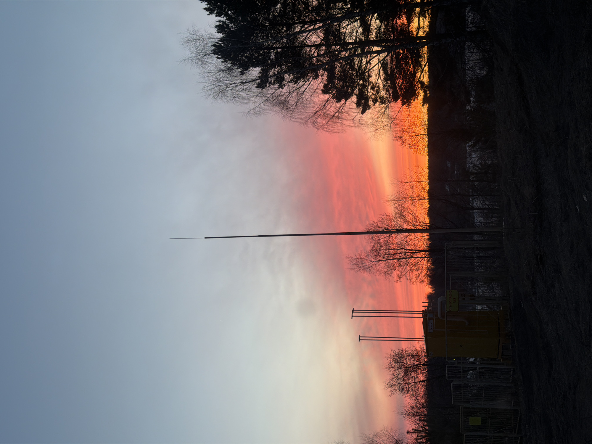 The image shows a sunset in a location near Izhevsk, Russia. The sky is divided into two sections. The left side is a pale blue, blending into a mixture of orange and pink towards the center. The right side is a vibrant orange and red. Silhouetted trees are visible along the top of the frame. A tall, thin pole extends upward from the center of the frame. Closer to the bottom, there is a yellow structure with fencing around it. The foreground is dark.
