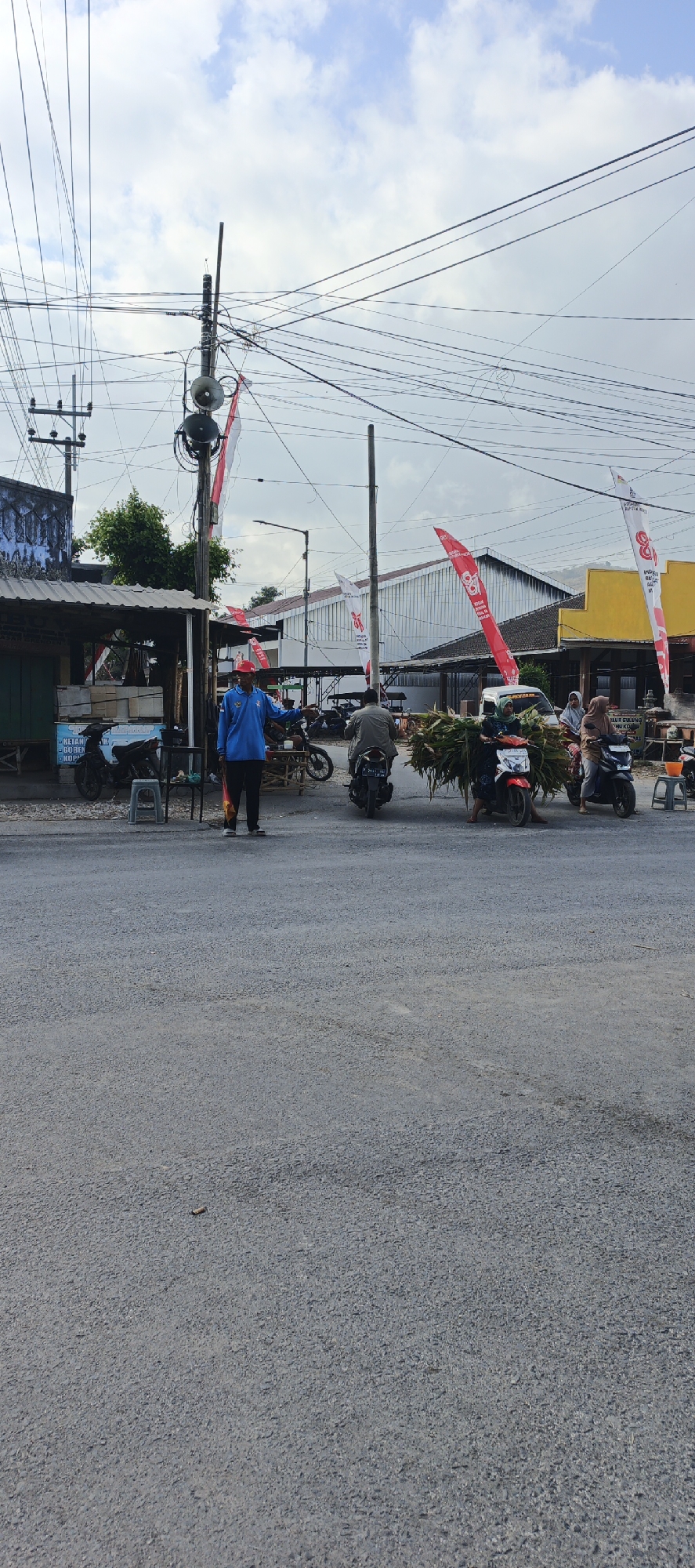 The image depicts a street scene in Grenden, Indonesia, under a partly cloudy sky. In the foreground, a man in a blue shirt and red cap stands in the middle of the asphalt road, holding a red object. He appears to be directing traffic. Several motorcycles are visible. One motorcycle, to the left of the man, is parked. To the right of the man, another motorcycle carries a large bundle of green foliage. Further to the right, a third motorcycle is also visible. Along the left side of the road, there is a small building or stall with a corrugated metal roof, and several motorcycles are parked in front of it. Red and white flags are displayed on poles throughout the scene. The background shows more buildings and a line of trees. Electrical wires are strung across the sky, crisscrossing overhead.