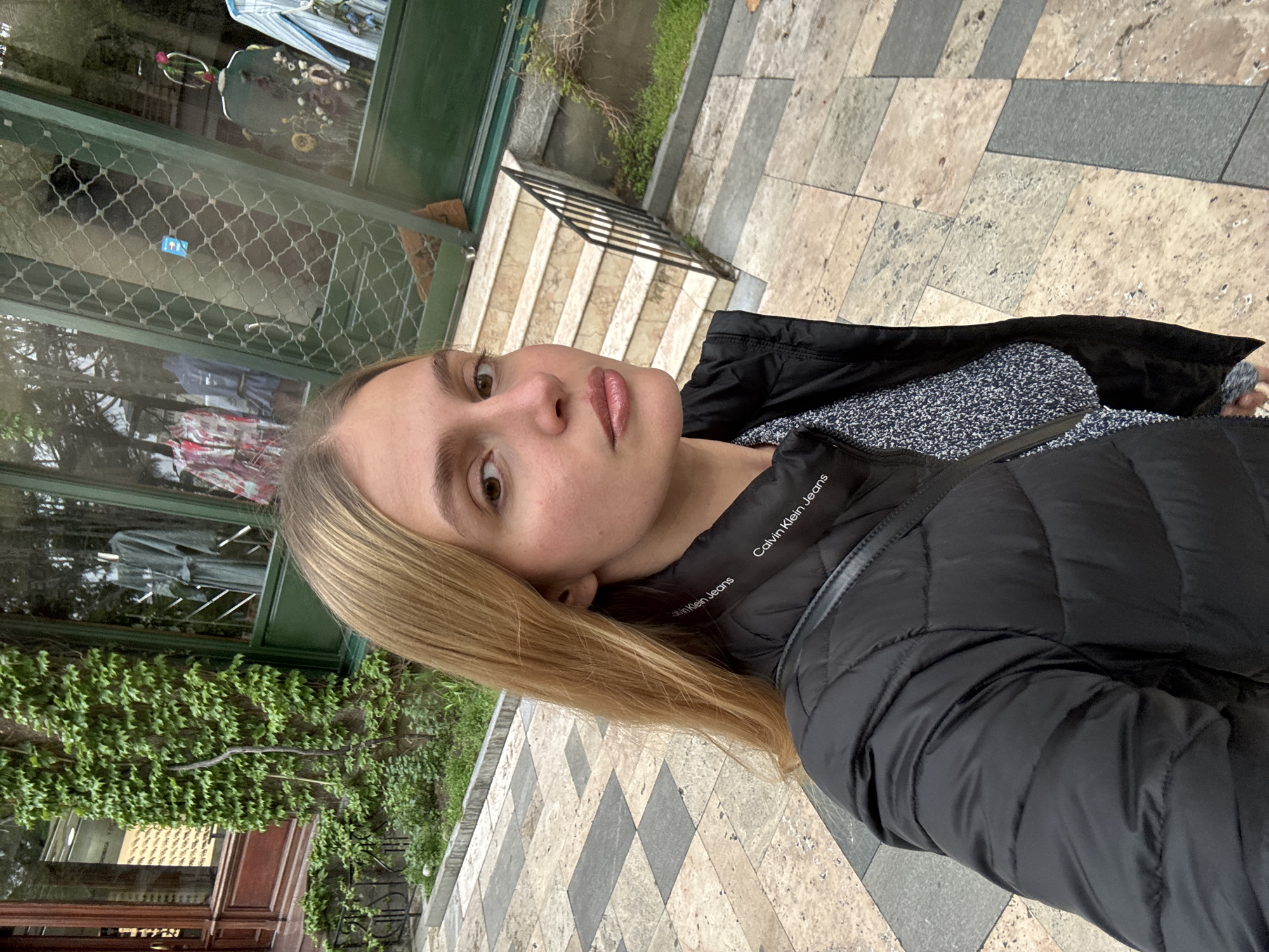 A young woman takes a selfie outdoors on a geometrically patterned stone walkway. She wears a black Calvin Klein Jeans puffer jacket over a blue and white patterned sweater. In the background, there is a building with a green-framed window covered in climbing ivy, a staircase with black railings, and additional greenery. The location is in an urban environment, potentially a sidewalk or pedestrian area.