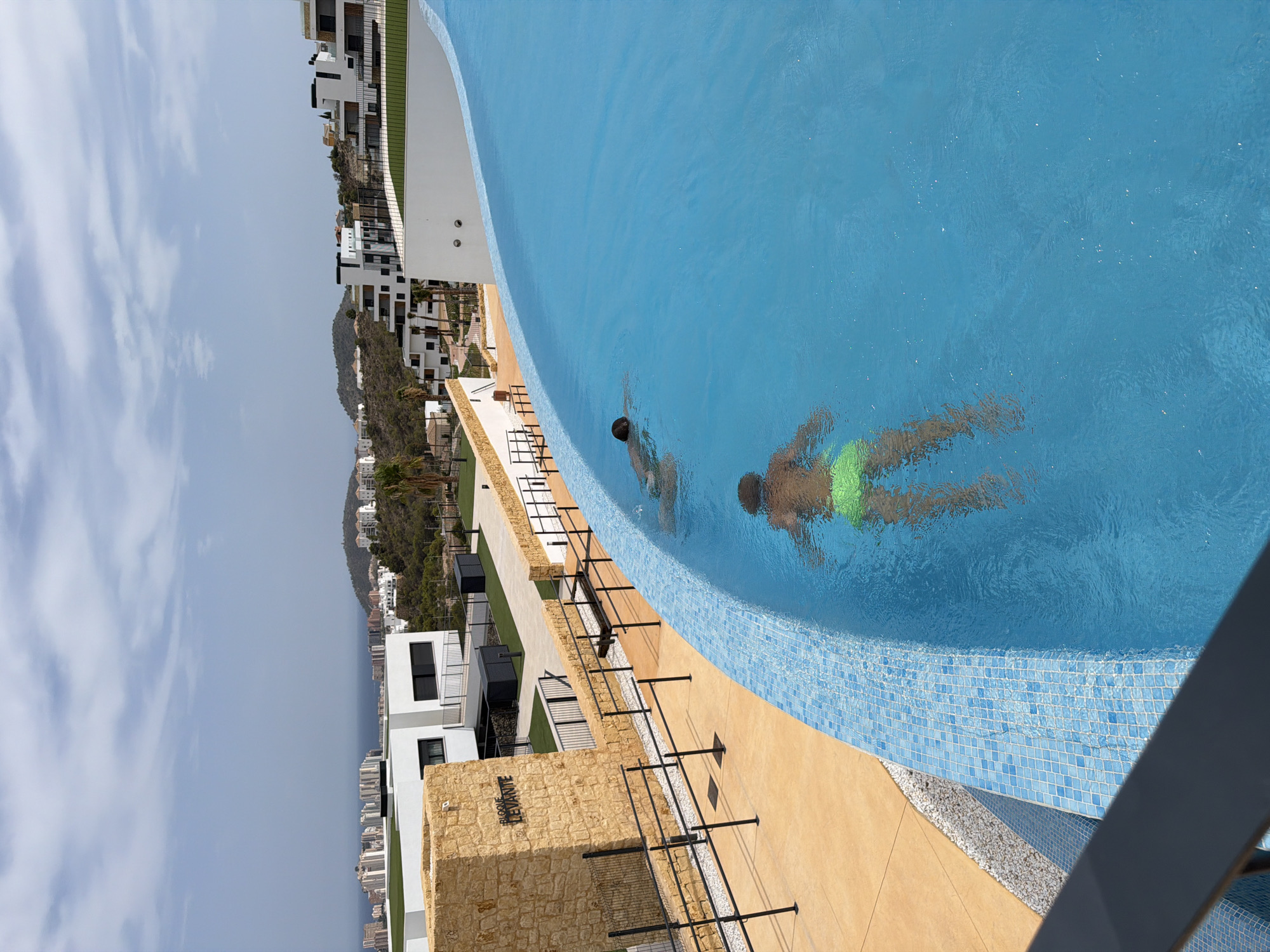 This is a vertical, eye-level, outdoor shot taken at a rooftop swimming pool in Finestrat, Spain. The pool's water is a vibrant blue, with a tiled edge. Two individuals are swimming in the pool, one in the foreground wearing green swim trunks, and another further back. The background shows a panoramic view of the city, including buildings, hills, and the sea under a cloudy sky. The area around the pool includes a tan-colored walkway with a textured surface, bordered by a low wall and a black railing. To the left, a stone structure with the inscription "BLOQUE LEVANTE" is visible, accompanied by more railings and gravel. The overall scene depicts a leisure setting within a developed urban and coastal environment.