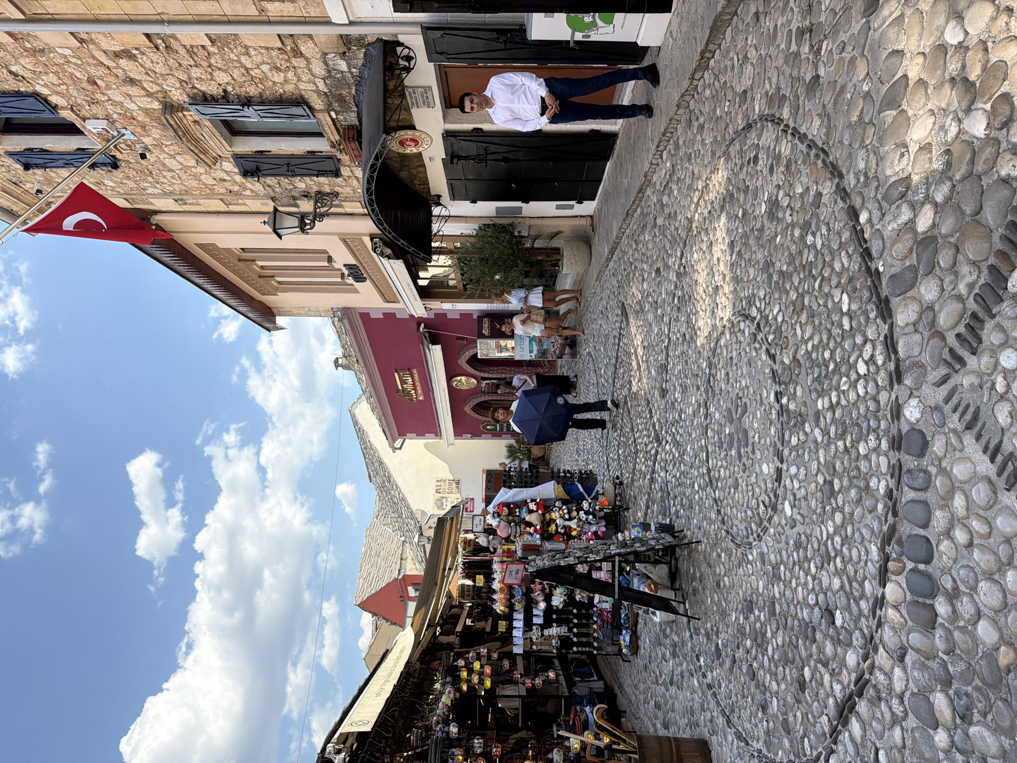 This media file captures a street scene in Mostar, Bosnia and Herzegovina, likely during daytime given the bright sunlight casting shadows. The image is oriented vertically, with the sky and buildings on the left and a cobblestone street on the right.
The foreground features a vibrant market stall overflowing with various souvenirs, including plush toys, decorative items, and possibly lamps. A person holding a dark umbrella stands near the stall. Further down the street, a man in a white shirt and dark pants poses for the camera. Other individuals are visible in the background, some walking and others gathered near buildings.
The architecture is characteristic of Mostar, with stone buildings, traditional shutters, and a Turkish flag prominently displayed on a flagpole. The street itself is paved with smooth, round cobblestones, some arranged in decorative patterns. The sky is a bright blue with scattered white clouds, suggesting pleasant weather. The overall atmosphere appears lively and tourist-oriented.
Visible text includes "lodije Stoga Grada Stanos," "ALL AVE SOME SOME GAVE ALL," and "LONDEUGHT & BAKLAV," "olloman," and "urban," which are likely names of shops or establishments. The scene conveys a sense of cultural tourism and local commerce.