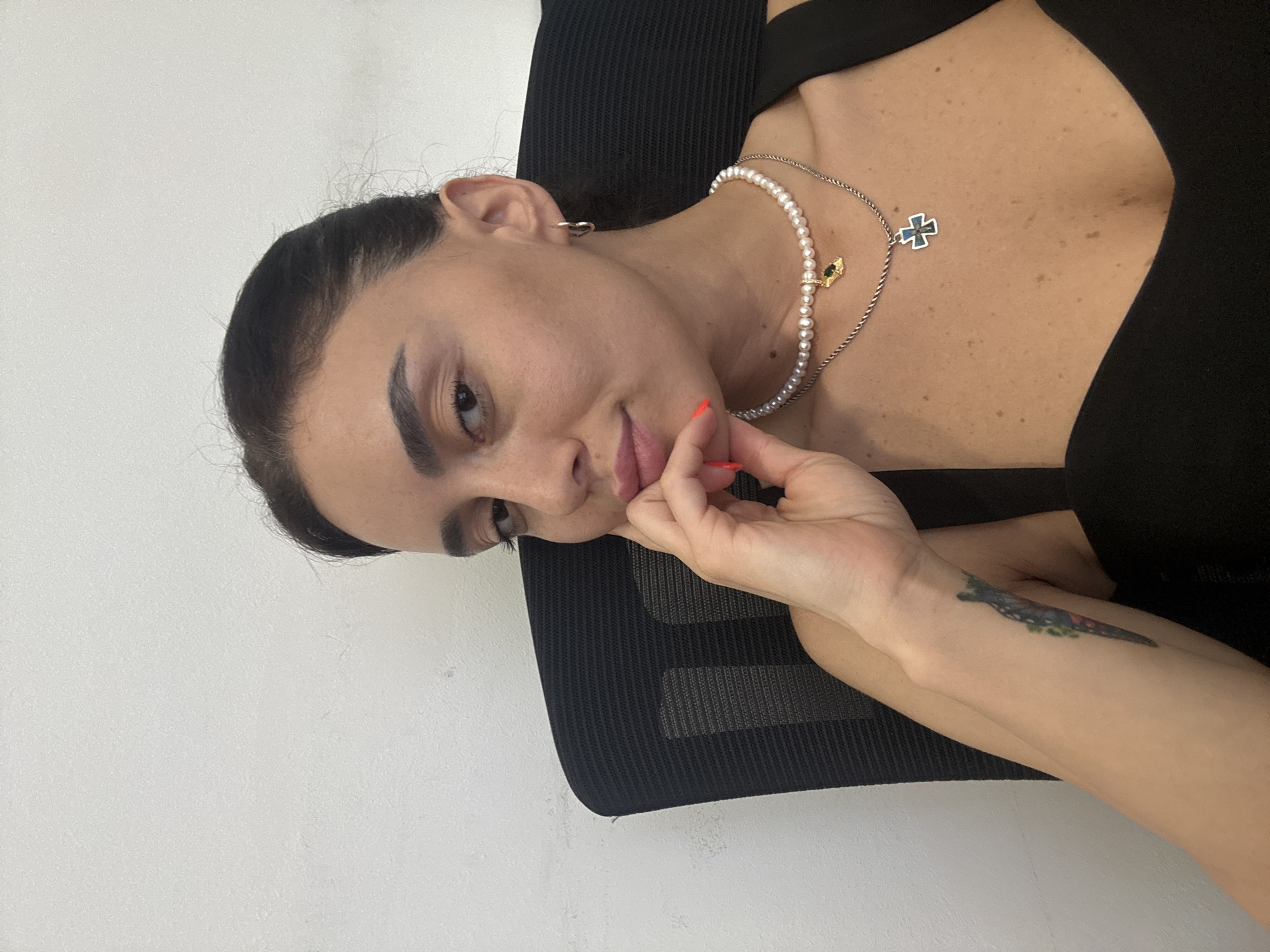 A young woman with dark hair and a dark top sits in an office chair in front of a white wall. She has her hand on her chin and is looking towards the camera. She is wearing a pearl necklace with a gold charm and a silver chain with a cross pendant. She has a tattoo on her left forearm. The image appears to have been taken indoors in a brightly lit room. The setting is likely an office or home office in Tbilisi, Georgia.