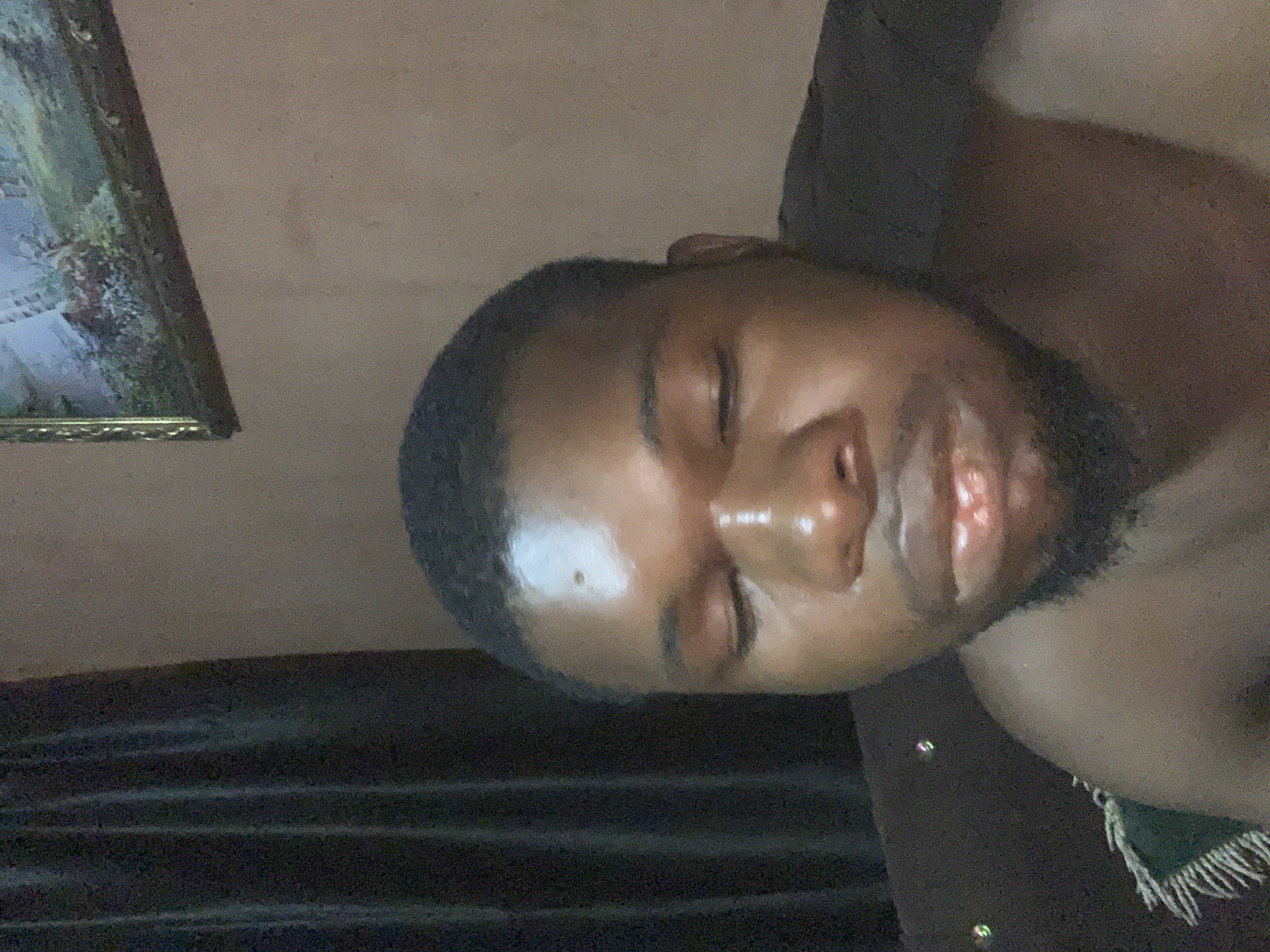 The image captures a close-up of a young man resting indoors. His head and part of his bare shoulder are visible, with his face angled towards the upper right. He has dark skin, short dark hair, and a beard or stubble. His eyes are closed or partially closed, indicating he is sleeping or deeply relaxed, and his facial skin appears slightly shiny.
The background suggests an indoor setting, likely a room. In the upper left corner, a framed picture or painting hangs on a light-colored, possibly beige or tan, wall. The artwork within the ornate golden frame appears to depict a landscape with greenery. The man is positioned against what seems to be a dark fabric, possibly a sofa or bed covering. On the right side of the frame, white fringes attached to a green fabric, possibly a blanket or throw, are visible.
The overall lighting is dim, suggesting it could be evening, night, or a softly lit interior during the day. The man's relaxed posture and closed eyes convey a peaceful emotion. There is no visible text within the image. The provided context indicates the location is Obosi, Nigeria.