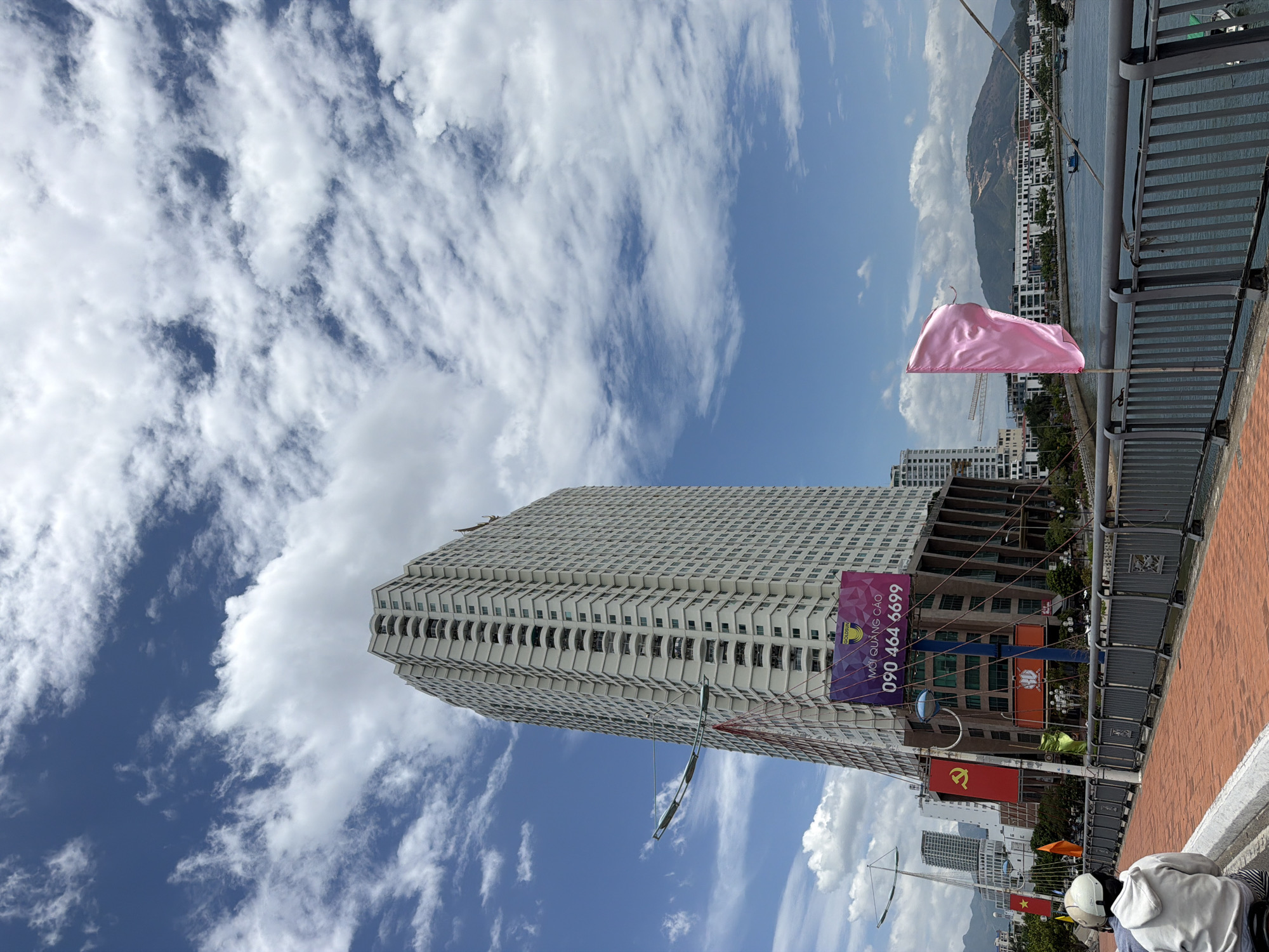A high-rise building with a white facade and many balconies stands tall against a blue sky filled with scattered white clouds. In the foreground, a person wearing a white hooded sweatshirt and a white helmet is seated on a scooter, facing away from the camera. They are on a walkway paved with red bricks, bordered by a gray metal fence. To the right of the walkway, a body of water, likely a river or bay, is visible, with several boats and buildings lining the opposite shore. A pink flag flutters on a pole to the right of the walkway. A large billboard with Vietnamese text and a phone number is attached to the lower part of the tall building. The scene appears to be in Nha Trang, Vietnam, a coastal city known for its beaches and high-rise developments.