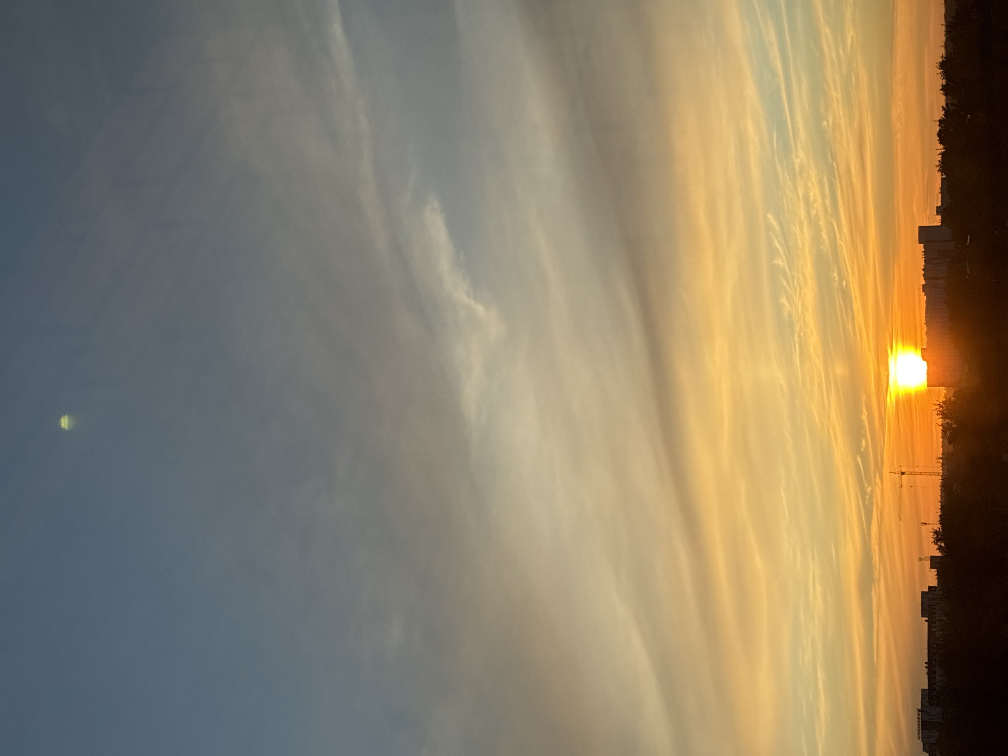 The image depicts a skyline at dusk in the city of Unknown, Unknown. The sky transitions from a pale blue at the top to a vibrant orange and yellow near the horizon, with thin, wispy clouds scattered throughout. A bright, setting sun is visible on the right side of the image, casting a warm glow. Silhouetted against the colorful sky are various buildings, including what appears to be apartment complexes and a construction crane. Trees and other foliage form a dark, dense line along the bottom of the frame, obscuring the ground. The overall impression is a serene, urban landscape at the end of the day.