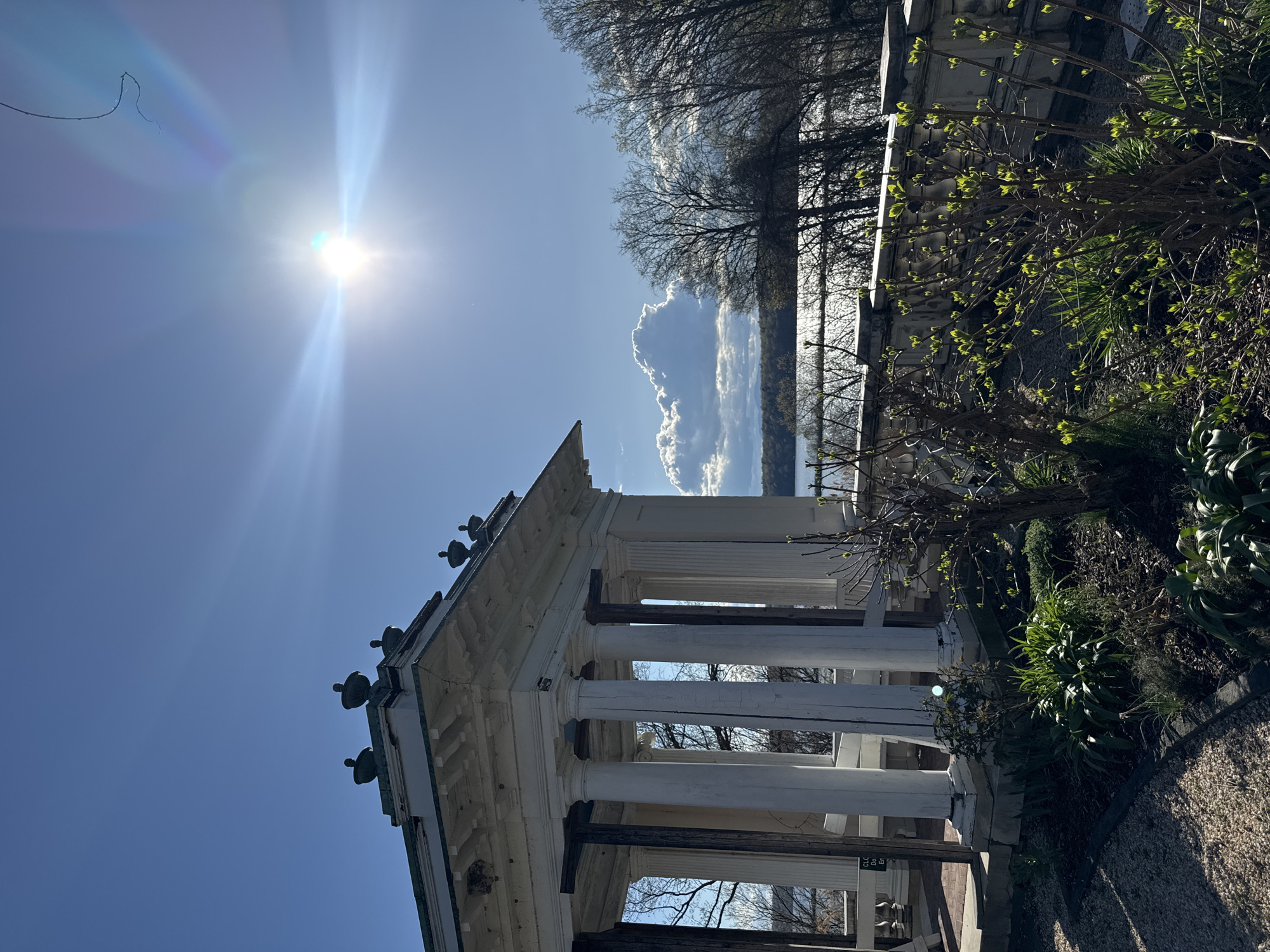 The image shows an outdoor scene featuring a white gazebo-like structure with classical columns, situated near foliage and trees. The sky is clear blue, with the sun shining brightly, creating lens flare in the upper left. A cloud is visible in the background. The gazebo is likely located in a park or garden setting. The image was taken in Town of Red Hook, United States of America.