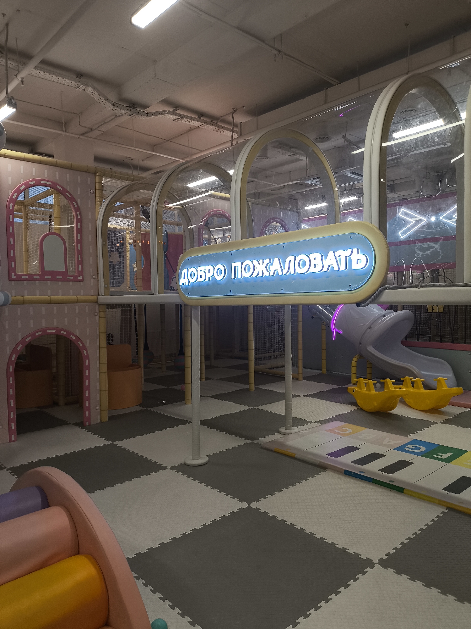 The image shows an indoor playground with soft, padded flooring and colorful play structures. A prominent feature is a blue neon sign that reads "ДОБРО ПОЖАЛОВАТЬ" (Welcome) in Cyrillic letters. To the right of the sign, a gray slide with yellow supports is visible, along with two yellow rocking toys. A musical keyboard-themed mat is laid out on the floor in front of the slide. The playground appears to be designed for children, with soft materials and engaging play elements. The lighting suggests an indoor setting, possibly a dedicated play center or entertainment facility. The overall impression is of a safe and fun environment for children. The context provided indicates this location is in Kaliningrad, Russia, but this information cannot be visually verified from the image alone.