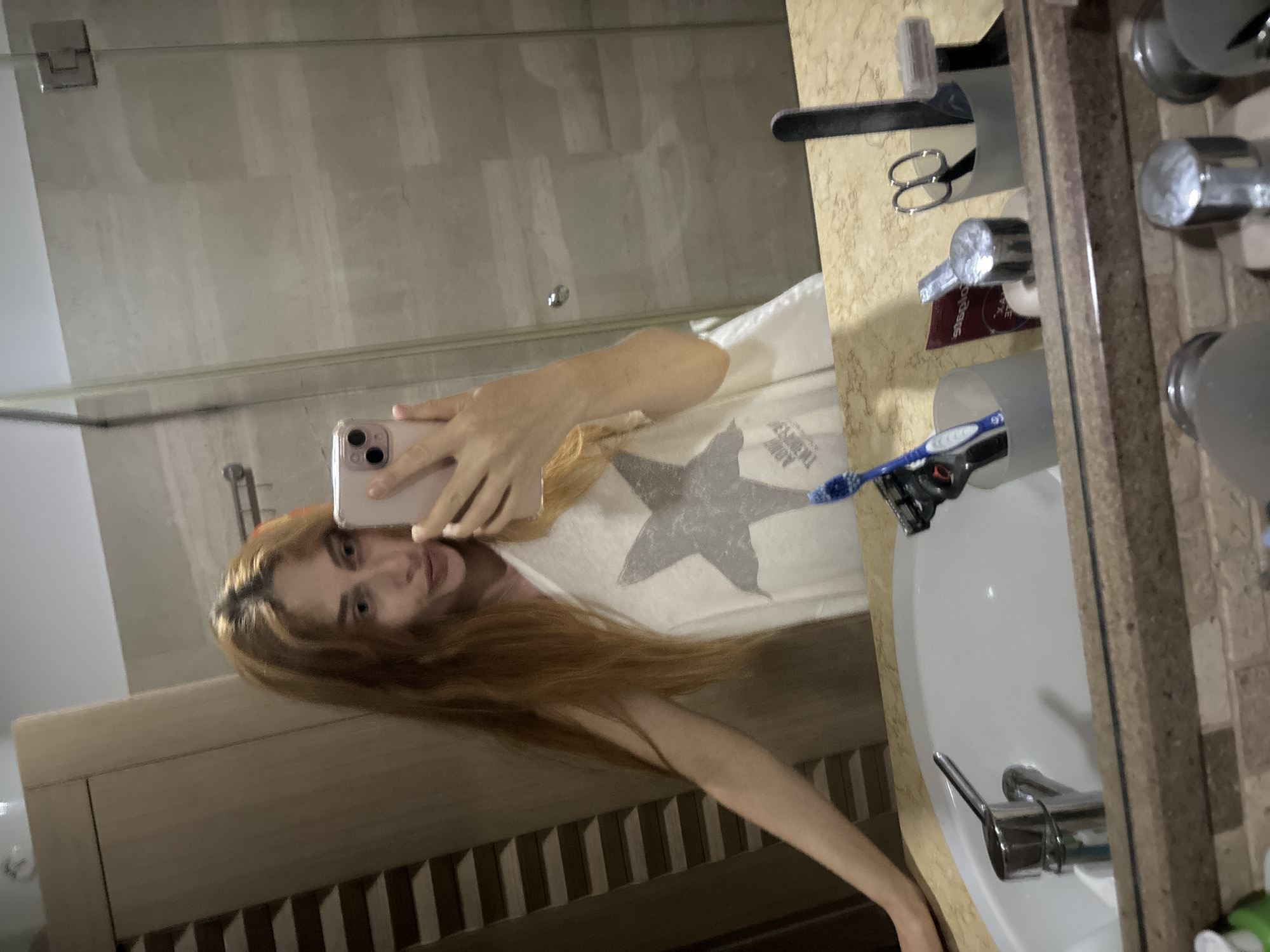 A young woman with long, auburn hair and fair skin takes a mirror selfie in a bathroom. She is wearing a white t-shirt with a large, grey star design on it, and is holding a pink smartphone in her right hand. Her left arm is resting on the edge of a marble-topped sink. The bathroom features a light-colored tiled wall, a glass shower enclosure, and wooden cabinetry. On the sink counter, visible in the mirror's reflection, are a faucet, a toothbrush, a razor, scissors, and several toiletry items. The overall impression is that of a private, domestic space, likely a hotel bathroom in Budva, Montenegro, given the provided context.