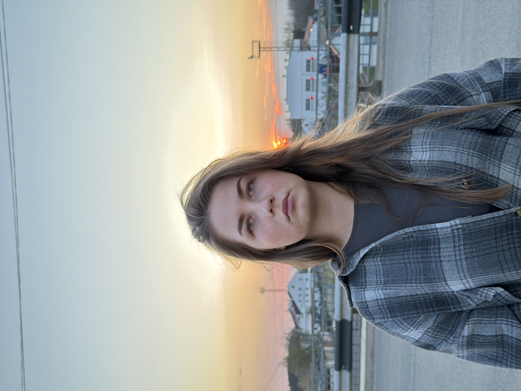The image shows a young woman with light brown hair and blue eyes, wearing a gray t-shirt and a gray plaid shirt. She is facing the camera with a neutral expression. The background features a sunset, infrastructure, and buildings. Based on the buildings and infrastructure, the location is likely the city of Chaikovsky, Russia. There are power lines and industrial buildings in the background, suggesting a semi-urban or industrial area. The photo was likely taken outdoors during the evening.