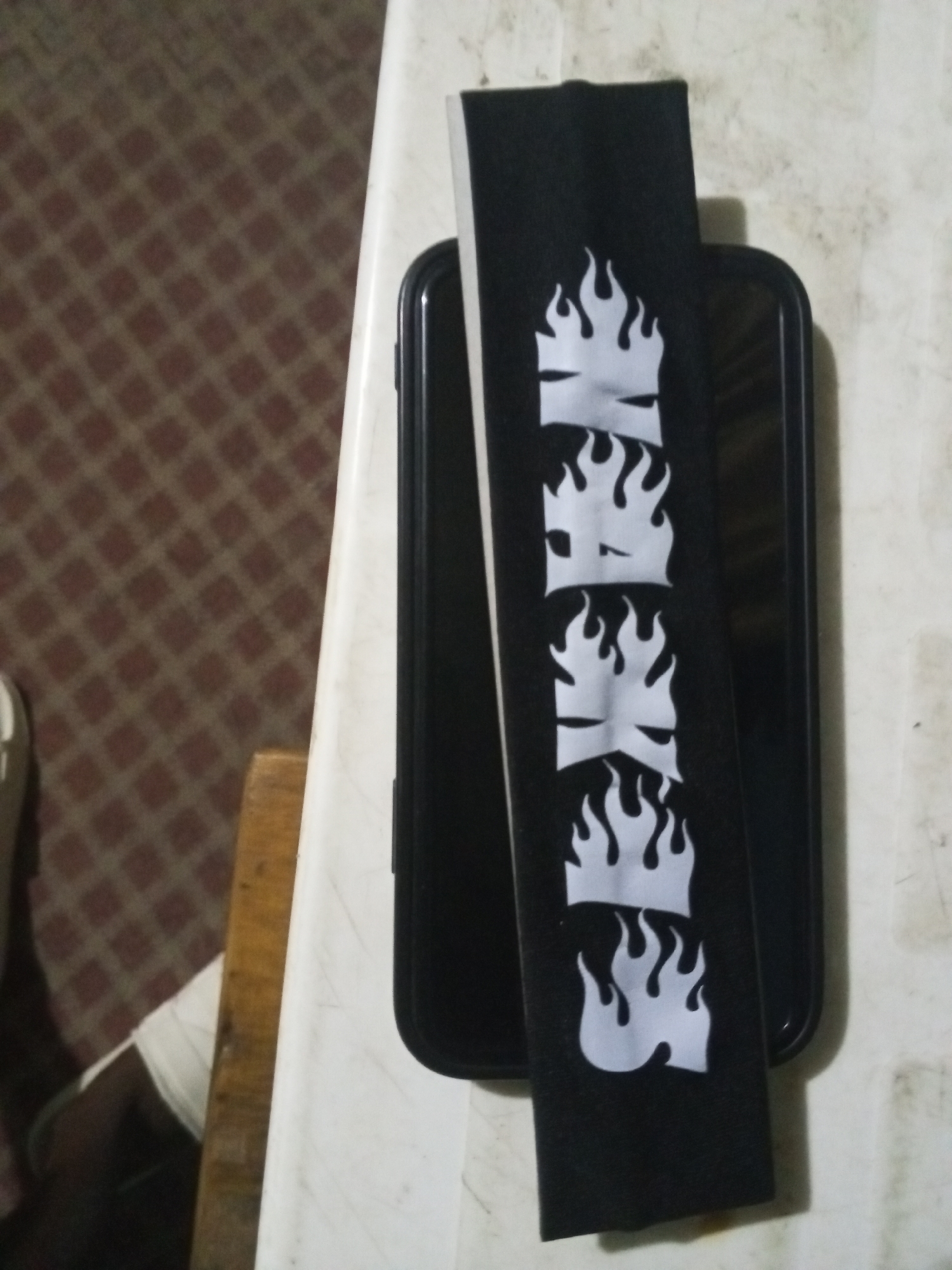 A black smartphone lies flat on a white surface, with a black armband displaying the word "SKATE" spelled vertically using flame-style lettering placed directly on top of it. The background reveals a brown checkered carpet and part of a chair leg, implying the photograph was taken indoors.