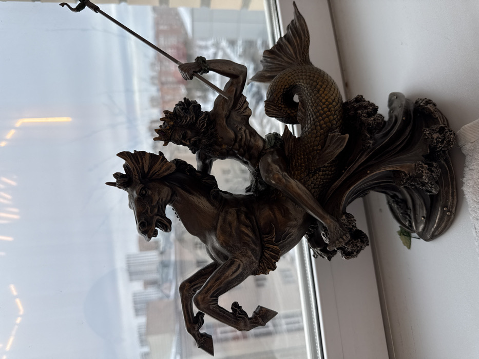 A bronze-colored statue depicts a male figure riding a sea horse-like creature emerging from waves. The male figure, possibly Poseidon, holds a long trident. The horse has a flowing mane and powerful legs. The statue is mounted on a wall or a windowsill, with a blurred urban landscape visible through the window behind it.
