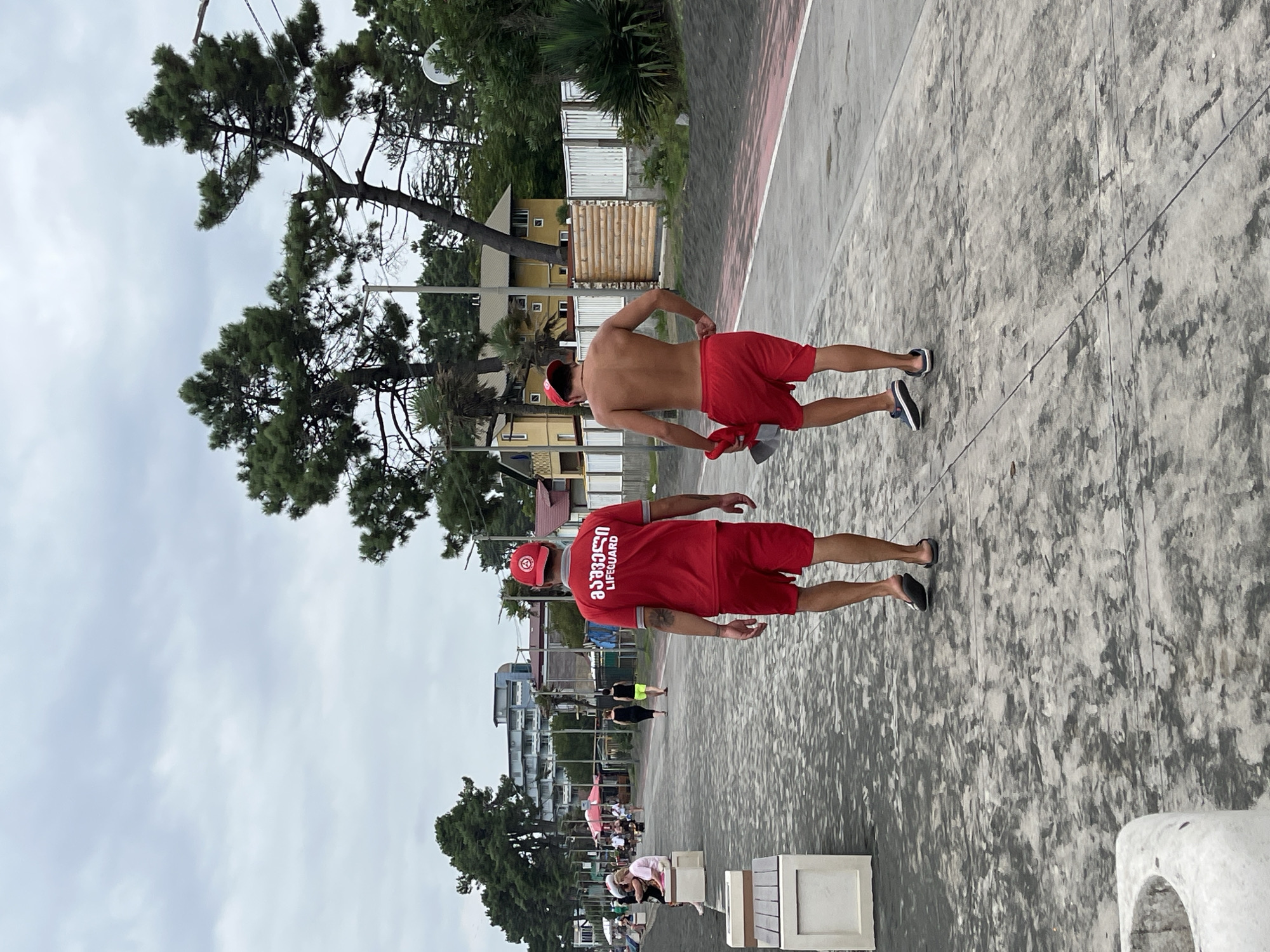 In Ureki, Georgia, two men in lifeguard attire walk along a paved promenade bordering a beach. The man on the left wears a red t-shirt with "LIFEGUARD" printed in white text and Georgian script, red shorts, and a red cap. The man on the right is shirtless, wearing red shorts and a red cap, and carries a red object in his left hand. The promenade is paved with a textured grey surface and has a red stripe along the right edge. In the background, a beach with dark sand is visible, lined with trees, buildings, and white fences. Several people are seen sitting on benches along the promenade to the left, and more figures are visible further down the beach and promenade. The sky is overcast with gray clouds.