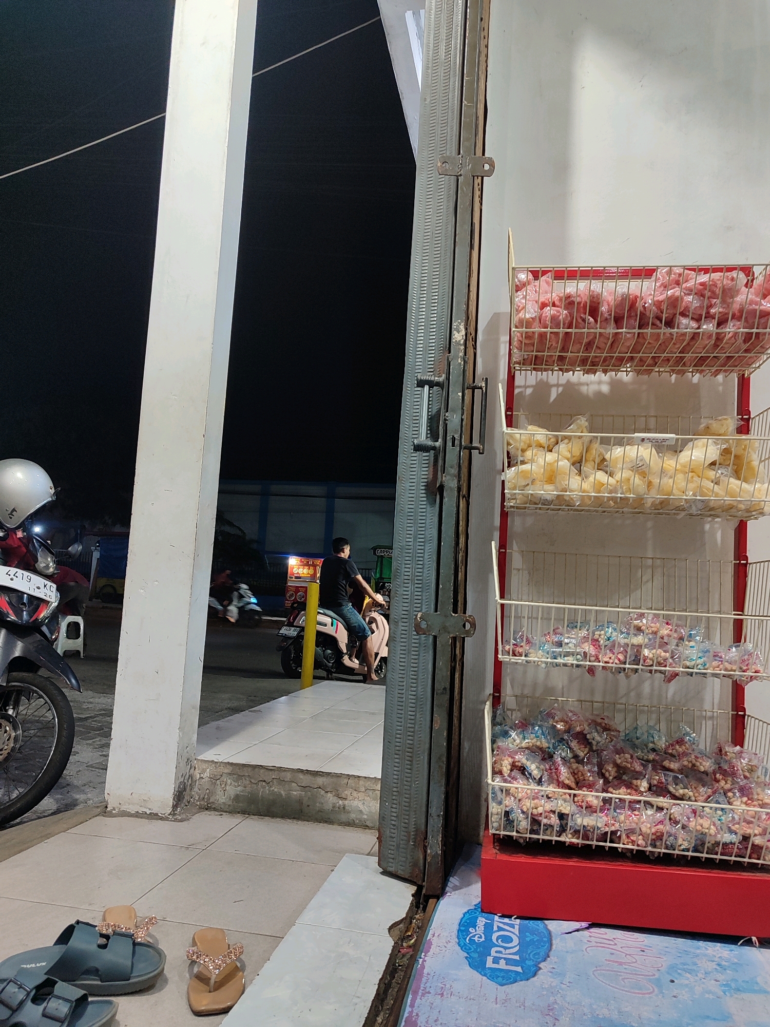 The image depicts an outdoor scene at night, likely a small storefront or roadside stall in Garut, Indonesia. In the foreground, there are several pairs of sandals on the tiled ground. To the right, a red display rack holds three white wire baskets filled with packaged snacks. The top basket contains pinkish items, the middle basket has light-colored snacks, and the bottom basket is filled with small, individually packaged nuts and candies. A portion of a blue and white patterned mat with "Disney Frozen" branding is visible on the lower right.
Behind the stall, a large white pillar is prominent, casting a shadow. Further in the background, a person is riding a light-colored scooter. A motorcycle with a silver helmet is parked to the left of the pillar. The overall environment suggests a street-side setting with ongoing activity, possibly a small business operating in the evening. The lighting is dim, indicating it's nighttime.