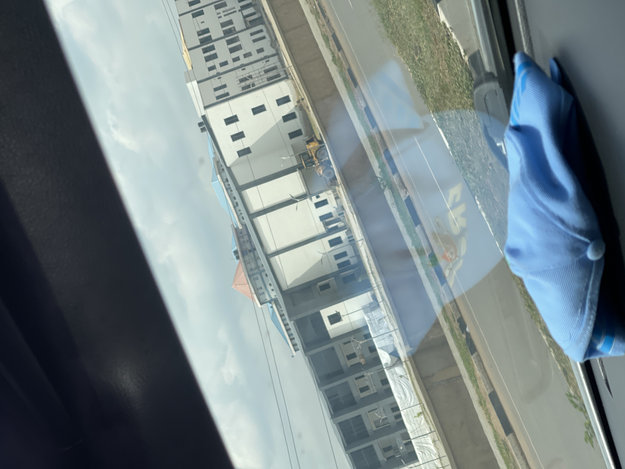 A low-angle shot through a vehicle window shows a view of Abuja, Nigeria. Prominent is a tall, modern building with multiple stories and a predominantly white facade with dark-framed windows. A section of road with a grassy shoulder runs parallel to the building. Reflected in the window is a blue baseball cap. There is also a blurred piece of construction equipment, such as a tractor or front-end loader, between the building and the road. The sky is overcast with scattered clouds.
