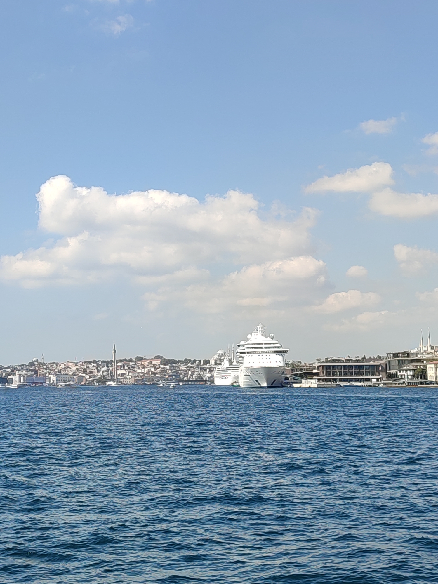 The image captures a wide shot of the Bosphorus Strait in Istanbul, Turkey, likely from a boat. In the foreground, the deep blue water of the strait is visible, with gentle ripples on its surface. Dominating the middle ground are two large white cruise ships docked or anchored. The larger ship is positioned closer to the viewer and its bow is prominently displayed. Behind the ships, the Beyoğlu district of Istanbul unfolds along the coastline. The cityscape is characterized by a dense collection of buildings with varying architectural styles, including some with distinctive domes and minarets, indicating a significant presence of religious or historical structures. A tall, slender minaret stands out amidst the urban landscape. The sky above is a bright blue, dotted with fluffy white clouds. The overall scene depicts a bustling port area with massive vessels juxtaposed against the historic architecture of a major city.