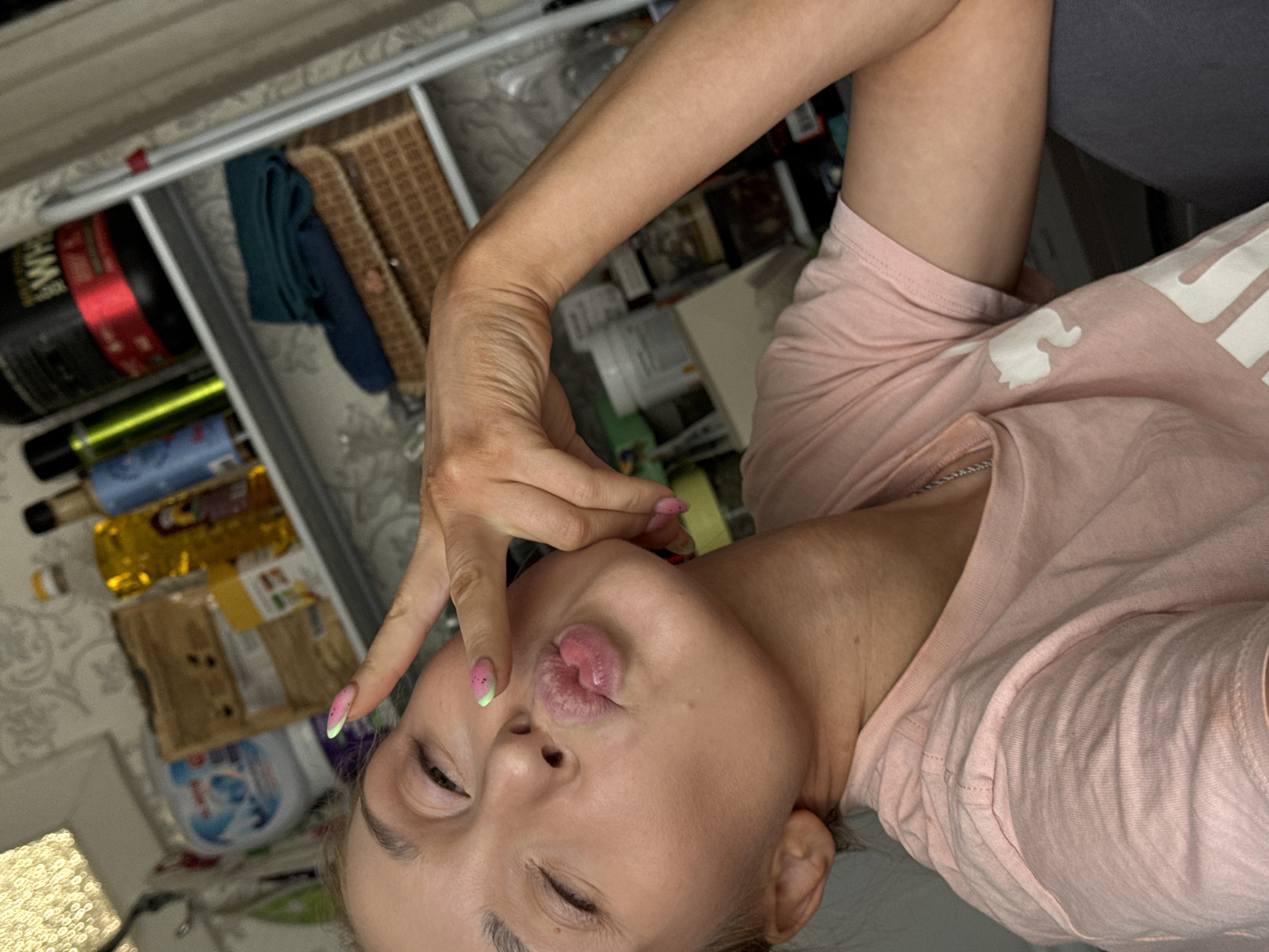A woman is lying on her back, wearing a pink t-shirt. She is holding her finger to her lips in a kissing gesture. The background includes a drawer with various items inside, including bottles and a woven basket. The location is the city of Chaikovsky, Russia.