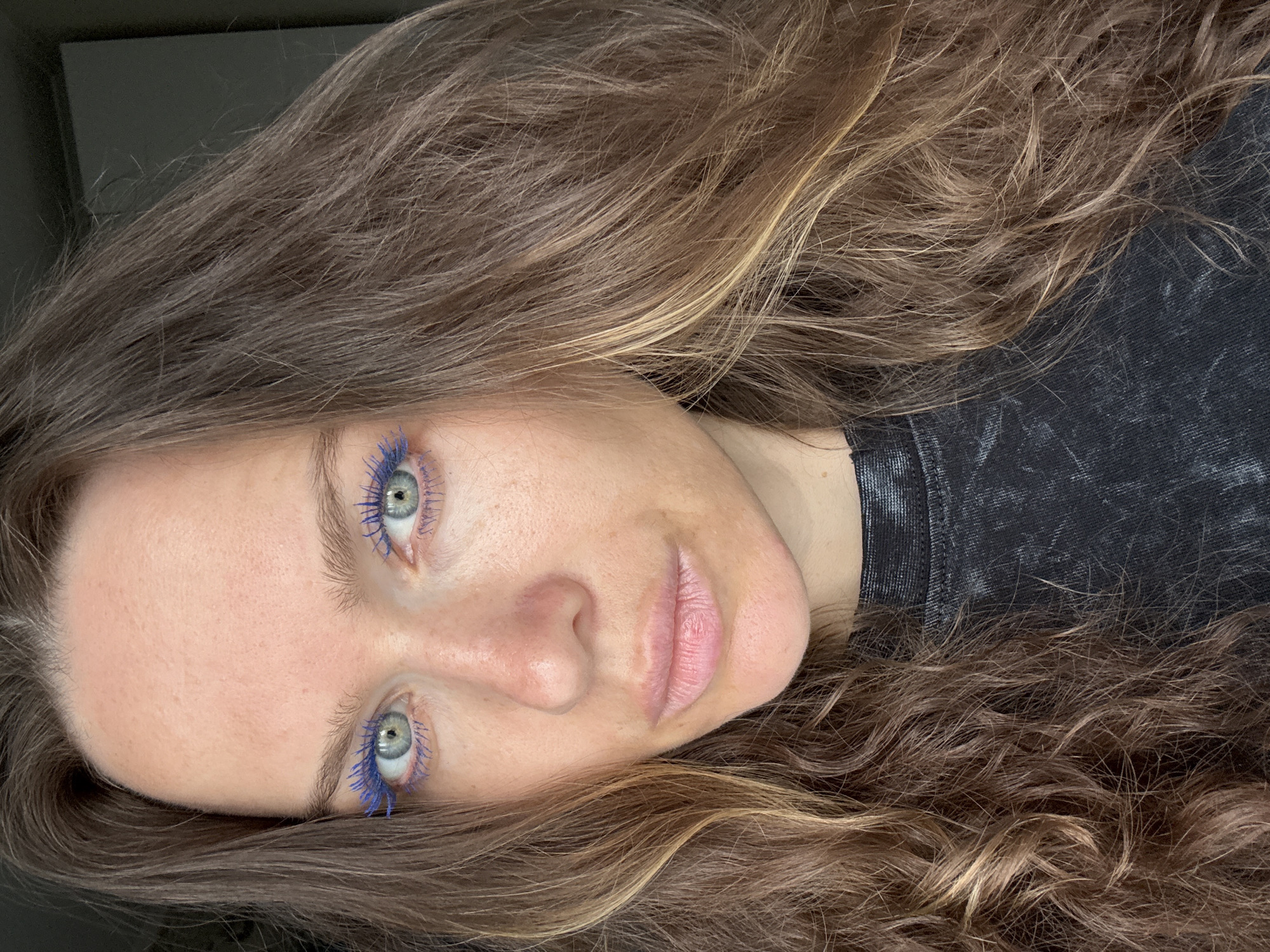 A close-up, slightly rotated portrait of a woman with light skin, green eyes, and brown hair with blonde highlights. Her eyelashes are coated with bright blue mascara. She is wearing a dark, possibly black, shirt with a textured or mottled pattern. The background is indistinct, appearing to be a dark, solid color. There are no signs of Moscow, Russia.