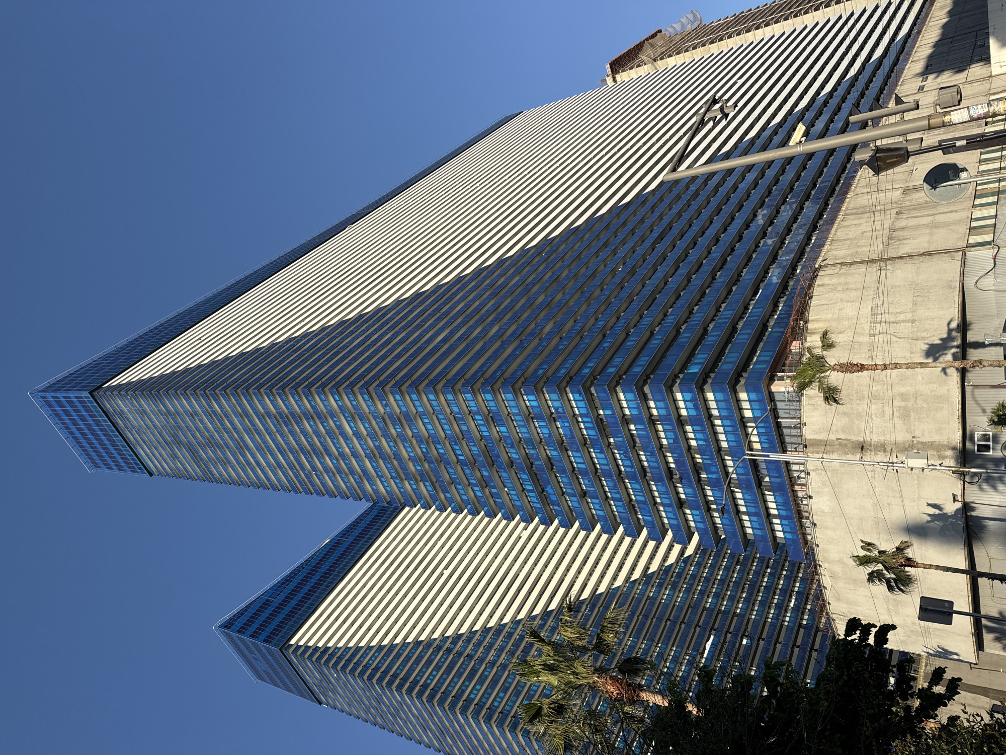The image depicts a low-angle view of two tall, modern buildings against a clear blue sky. The buildings are characterized by a repetitive pattern of horizontal white and blue lines, possibly reflecting sunlight. In the lower right of the image, portions of adjacent structures can be seen, including a street-level building with palm trees nearby and what appear to be streetlights and signs. The orientation of the image suggests it was taken looking upwards. These are the twin towers of Batumi, Georgia.