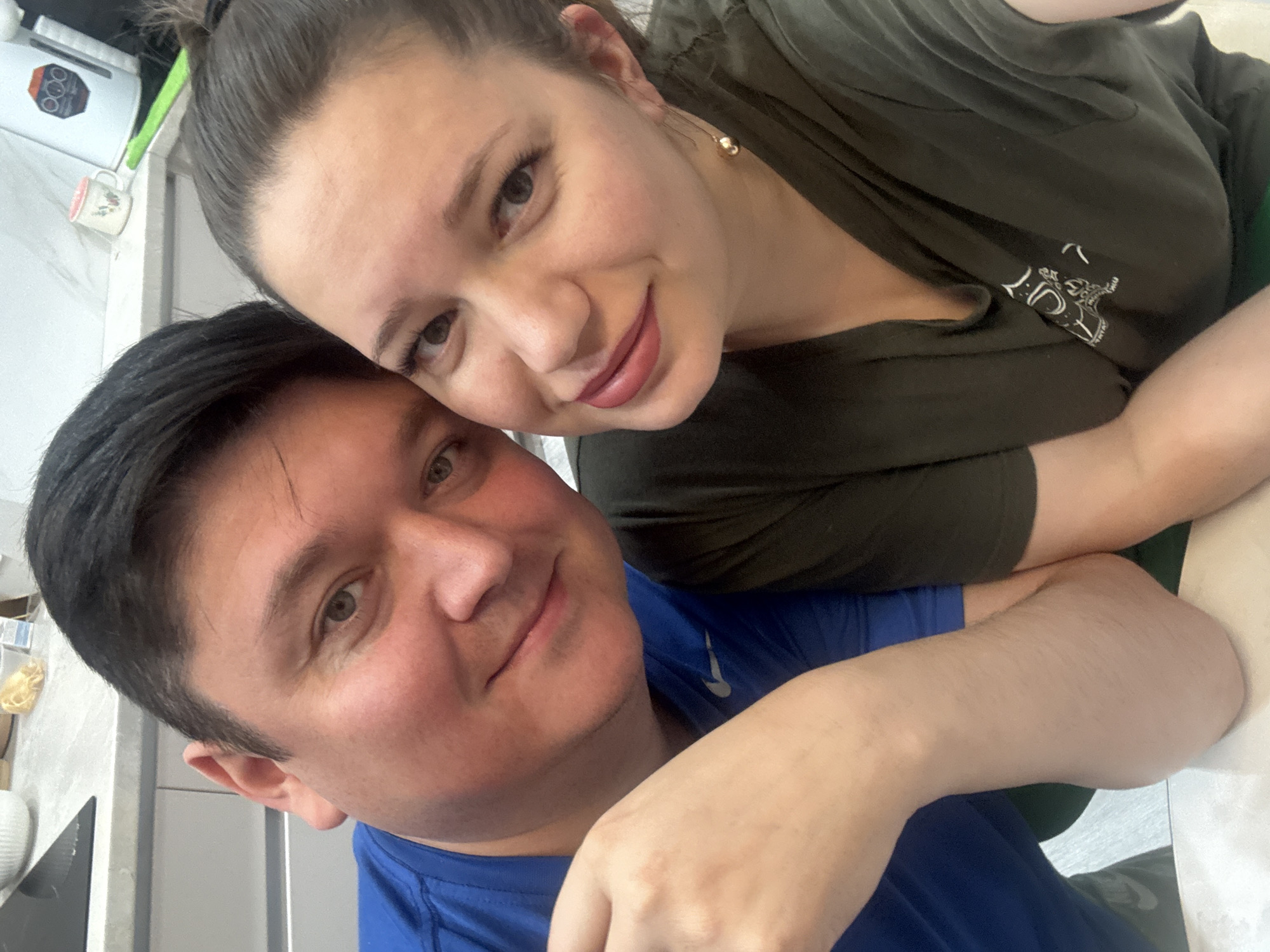 A close-up, rotated selfie features two adults, a man and a woman, posing intimately in an indoor setting, likely a home in Izhevsk, Russia. The woman, with dark hair pulled back and wearing a dark olive green t-shirt, leans on the man. She has a gentle smile, natural makeup with soft pink lipstick, and a golden, ball-shaped earring visible on her right ear. A white graphic design can be seen on the sleeve or shoulder of her shirt.
The man, with short dark hair, also smiles softly at the camera. He is wearing a blue t-shirt with a white logo, strongly resembling the Nike swoosh, visible on his right shoulder. Their expressions convey warmth, affection, and contentment.
In the background, slightly out of focus, is a light-colored counter or shelf. Objects visible include a white kitchen appliance with dials or buttons, and a white mug with a subtle pattern. The lighting is soft and consistent, indicative of an indoor environment. No specific time of day or weather is discernible. The overall scene suggests a happy, domestic moment between the two individuals.