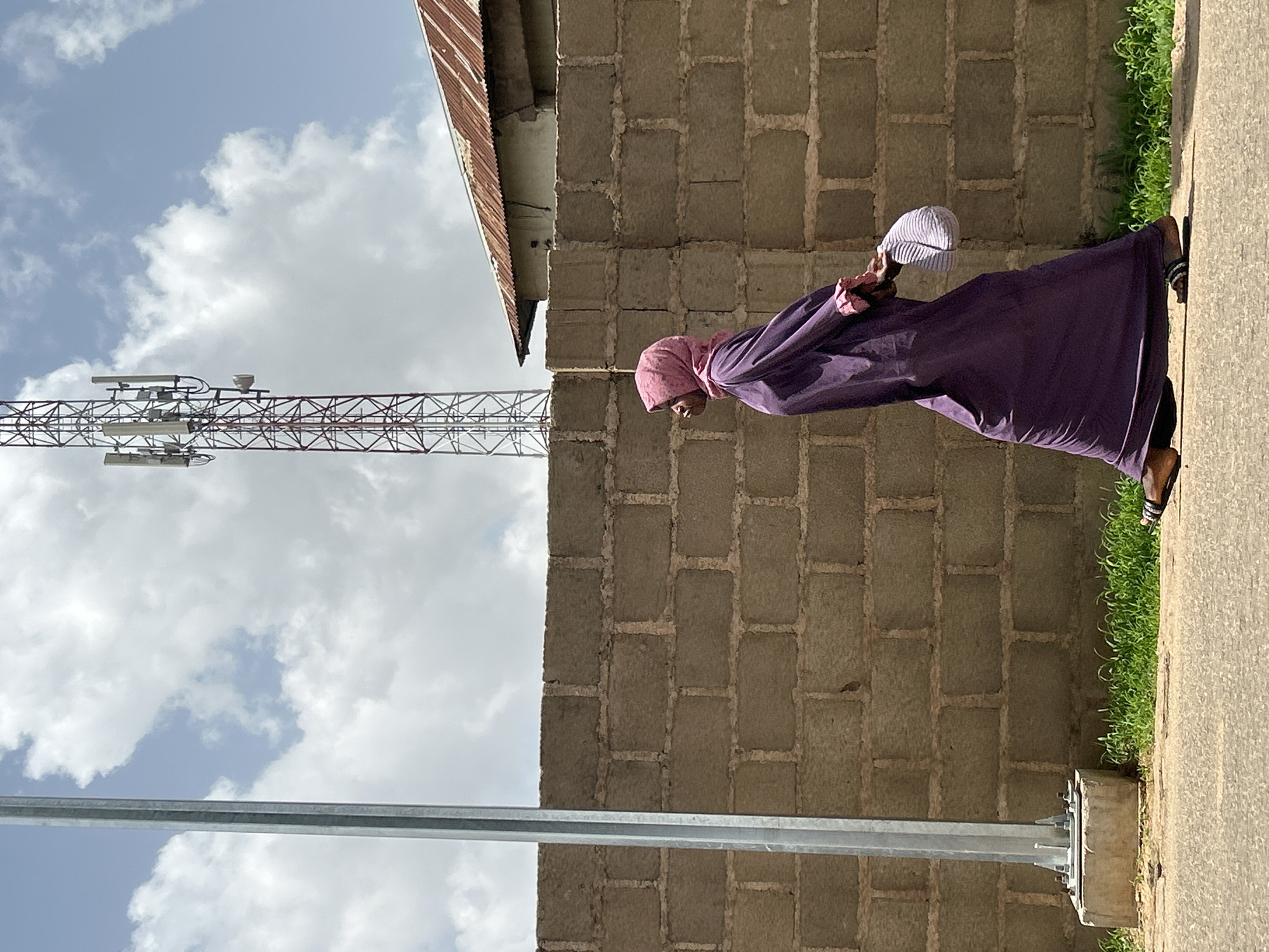 A woman in a pink hijab and purple dress walks along a sidewalk in Jos, Nigeria. She is holding a white hat in her hands. To her right is a brick wall with green grass growing at the base, and a rusted roof is visible at the top of the wall. To her left is a metal pole and a cell phone tower against a sky of white clouds.