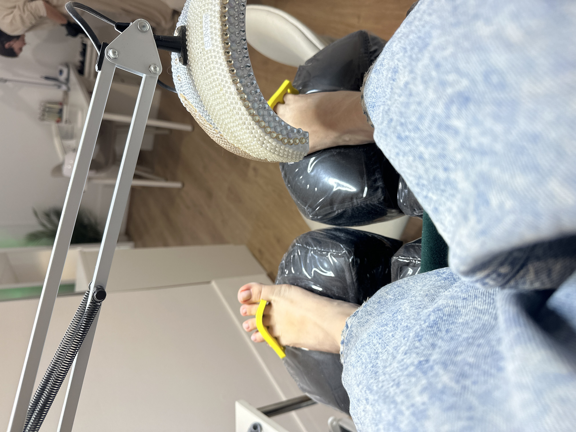 This eye-level, first-person perspective image depicts a person's feet undergoing a pedicure in an indoor setting, likely a nail salon. The person is wearing light blue denim jeans, visible on the right side of the frame. Their bare feet are placed on two black, plastic-covered footrests. Both feet have bright yellow toe separators positioned between the toes.
On the left side of the image, an articulated white desk lamp with visible springs and joints extends upwards. Next to the left foot, a "SUNone" brand UV/LED nail lamp is prominent. This lamp is distinctively embellished with numerous clear and gold-toned rhinestones or gems around its edge. The "SUNone" logo is clearly visible on its surface.
In the blurred background, reflections suggest the presence of another individual, possibly a nail technician, and various white furniture, shelves, and containers typical of a nail care environment. The floor appears to be light-colored wood. The overall scene indicates the ongoing process of a pedicure, possibly involving gel polish that requires curing under the UV/LED lamp. The atmosphere appears clean and well-lit. The specified location context is Zagreb, Croatia, although no specific visual cues in the image confirm this.
