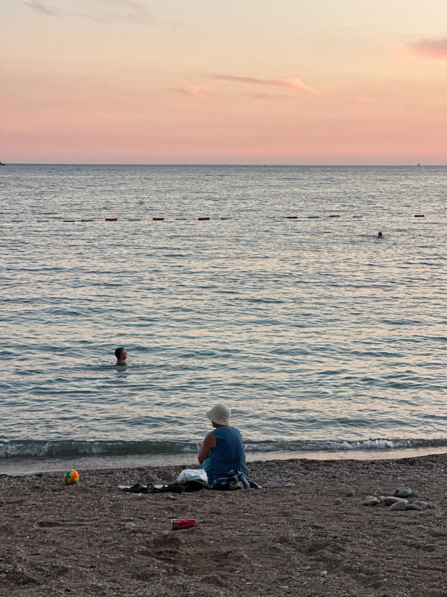 At sunset on the beach in Budva, Montenegro, a person sits on the shore facing the sea. The person is wearing a blue shirt and a white hat, and a beach ball and a drink can are visible on the sand near them. The sea is calm with gentle waves lapping at the shore, and in the distance, a single swimmer can be seen in the water. The sky is a soft pastel pink, indicating the end of the day.