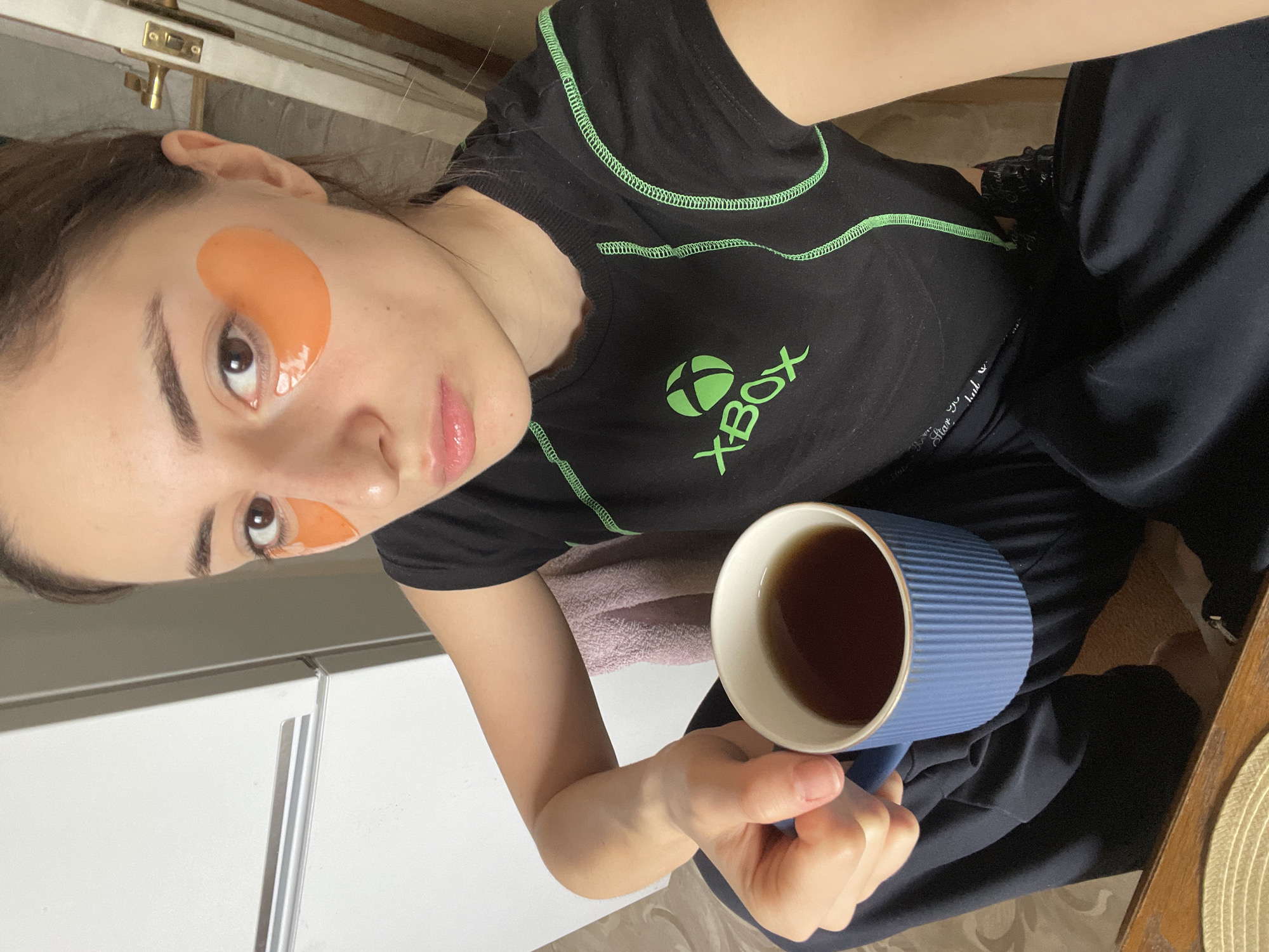 A young woman is captured in a high-angle, rotated selfie, looking directly at the camera with a neutral expression. She has dark hair, dark eyes, and a slight sheen on her lips. Orange, gel-like under-eye patches are applied beneath both eyes, suggesting a skincare routine or relaxation.
She is wearing a black t-shirt with a prominent green "XBOX" logo and text on the chest, along with decorative green stitching along the seams. Her lower body is clad in dark-colored, loose-fitting pants or tracksuit bottoms. In her right hand, she holds a ribbed, blue mug with a gold rim, containing a dark beverage, likely tea or coffee.
The setting appears to be an indoor, casual environment, possibly a home in Moscow, Russia, as per the provided context. In the background, white cabinet doors or furniture are visible, along with a glimpse of a wooden surface, potentially a table or chair, and a light-colored woven placemat. A small section of a pink or light-colored towel is also visible behind the mug. The overall scene conveys a relaxed, everyday moment, possibly during a morning or evening routine.