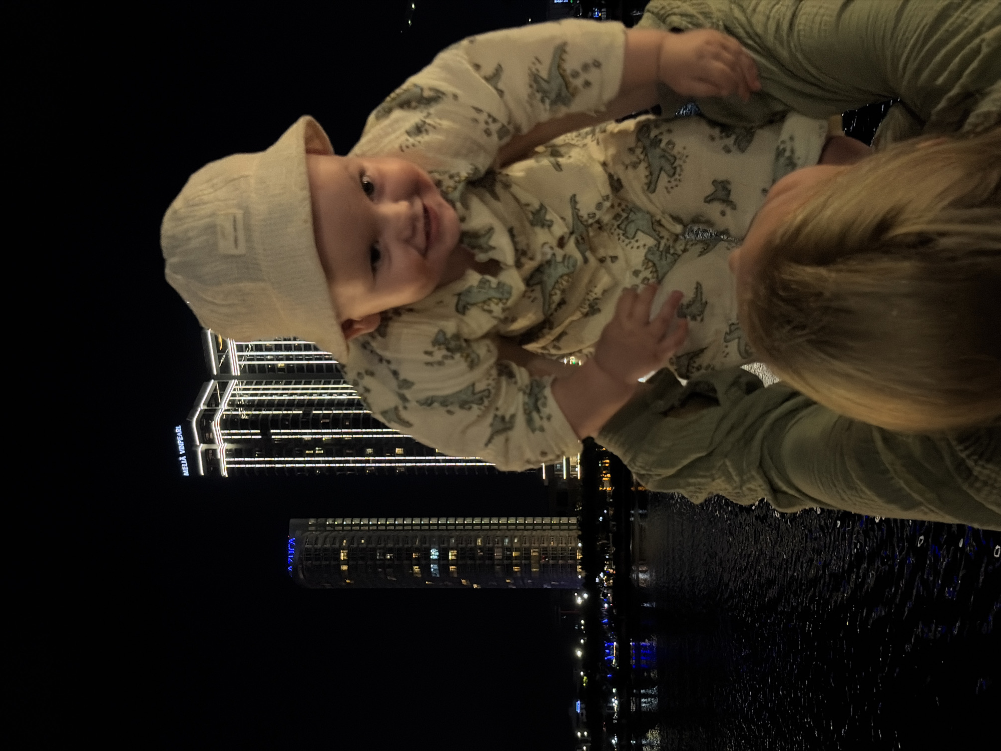 The image captures a heartwarming nighttime scene in Đà Nẵng, Vietnam, featuring a smiling baby held aloft by an adult against a backdrop of illuminated city skyscrapers and a reflective body of water.
The primary subject is a baby, looking directly at the camera with a wide, happy smile. The infant is wearing a light-colored bucket hat and a long-sleeved outfit patterned with what appear to be small dinosaurs. Only the top of the adult's head, with blonde hair, and a green-sleeved arm holding the baby are visible, suggesting an intimate interaction.
The setting is an urban waterfront at night. In the background, two prominent high-rise buildings glow with artificial lights. One building clearly displays the illuminated text "MELIÁ VINPEARL," while another, further away, shows blue-lit windows and partially visible text that appears to be "AZURA." A body of water, likely a river or bay, stretches across the bottom of the frame, reflecting the city lights and showing additional illuminations along its banks. The sky is dark and clear.
The overall impression is one of joy and affection, with the happy baby being the focal point against the vibrant, modern cityscape after dark.