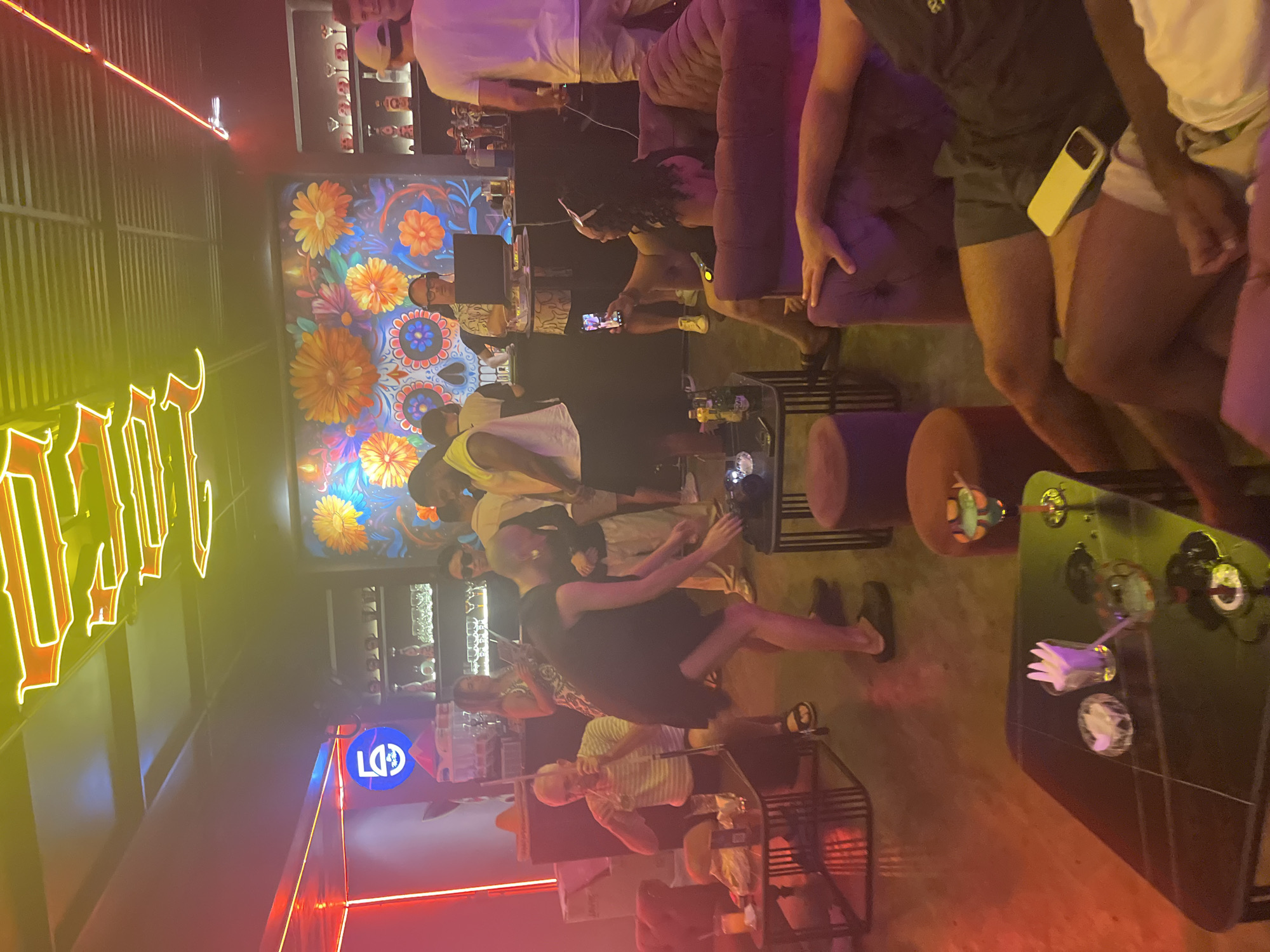 The image depicts a lively bar scene, potentially in Đà Nẵng, Vietnam. Several patrons are engaged in various activities. Some are seated at tables, while others are standing and interacting. There are tables with drinks and other items. The bar has a visually striking wall mural featuring colorful floral and skull motifs. Neon signs are prominent, along with other bar decor.