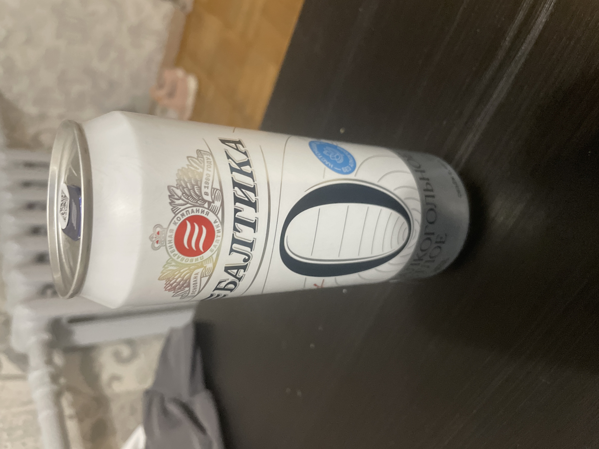 The image shows a can of Baltika beer lying horizontally on a dark, possibly wooden, surface. The can is predominantly white with blue and grey/silver accents. The brand name "БАЛТИКА" is prominently displayed in large, vertical letters. The can also features the Baltika logo with three wavy lines inside a red circle. The surroundings include a blurred background with what appears to be a piece of furniture with a patterned design. The photo does not contain information to place the beer can's location in Sarapul, Russia.