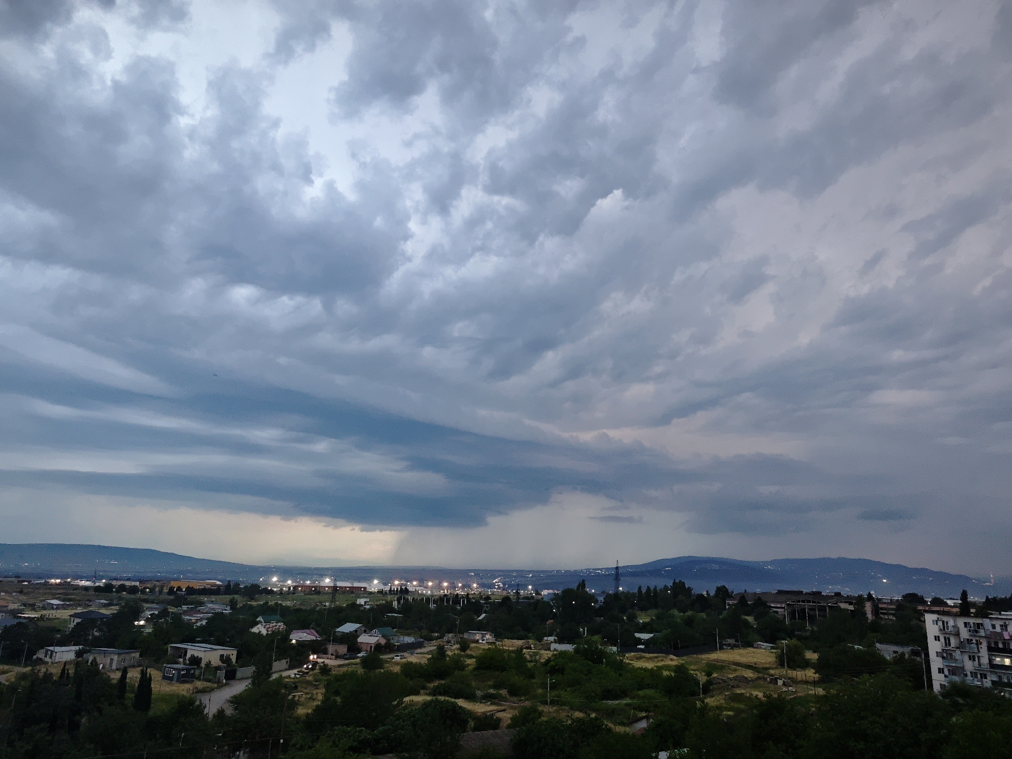 This is a wide-angle, high-angle photograph of a suburban area on the outskirts of Tbilisi, Georgia, taken from an elevated position, possibly a balcony or rooftop. The time of day appears to be dusk, indicated by the overcast sky and the artificial lights beginning to illuminate the distance.
The sky dominates the upper half of the frame, filled with heavy, gray, and blue clouds that suggest impending rain. There are streaks of lighter sky visible through gaps in the clouds, particularly in the upper right quadrant. In the far distance, beyond the urban sprawl, a range of low, dark mountains is visible.
The middle ground of the image depicts a suburban landscape with numerous houses of various sizes and architectural styles. Many of these houses have pitched roofs, some of which are brightly colored, adding small accents of red and green to the scene. The houses are nestled amongst lush, dark green trees and vegetation. Roads and pathways wind through the residential area.
Further in the distance, a more built-up area is visible, characterized by industrial buildings, factories, and possibly a power plant, indicated by illuminated structures and tall towers. A faint line of lights suggests a road or railway running through this area. The overall impression is that of a sprawling urban environment with distinct zones of residential and industrial development.
The foreground is also densely covered in dark green trees and foliage, obscuring the ground level. The light is dim, and details in the foreground are less distinct. There are no visible people or animals in the image. The scene conveys a sense of calm, perhaps before or after a rain shower, with the muted colors and the distant glow of city lights. The overall atmosphere is one of quiet observation of the urban and natural landscape.