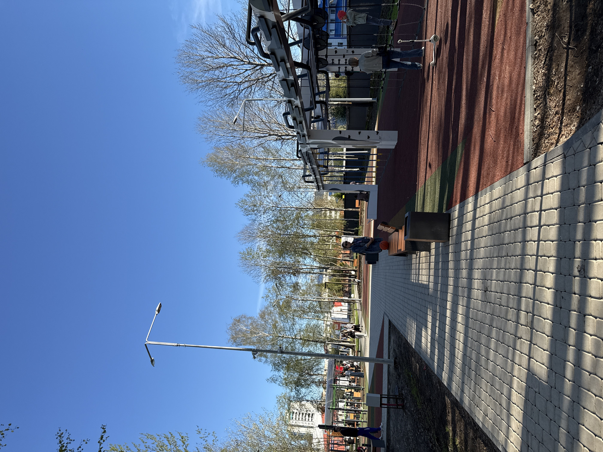 The image shows a public outdoor space in Tobolsk, Russia. A paved walkway made of gray interlocking bricks runs alongside a red athletic surface. Outdoor exercise equipment is visible, with people interacting with it. A man sits on a bench near the walkway. Streetlights are present along the walkway, and bare trees line the area. Buildings are visible in the background. The sky is clear and blue.