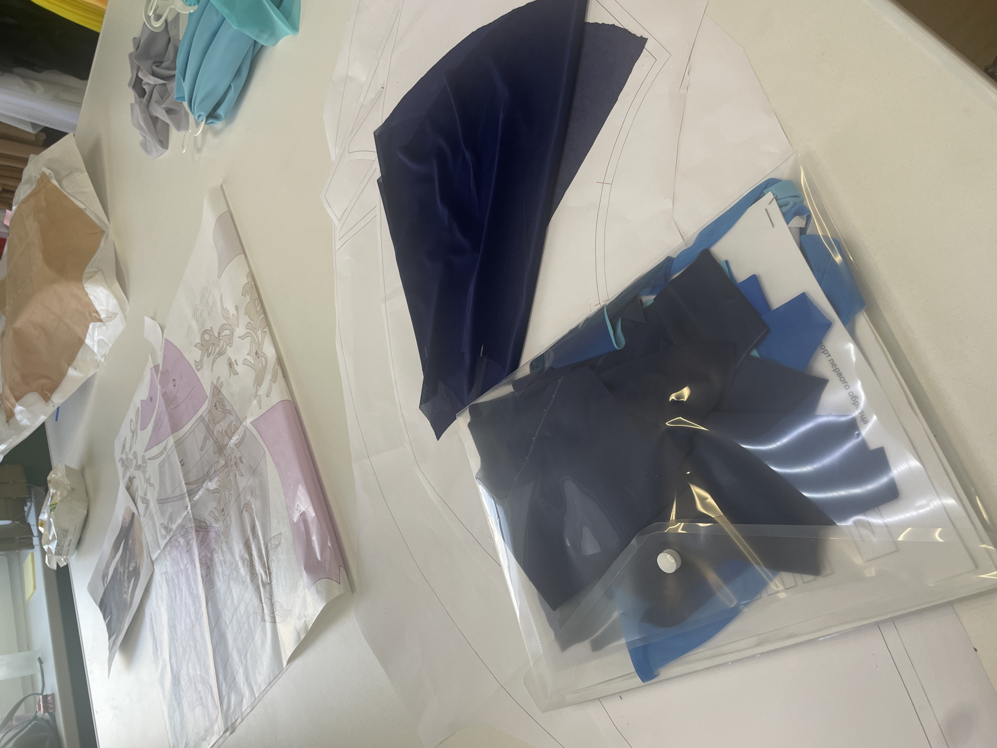 The image shows a white tabletop with various sewing supplies and fabric. In the foreground, a clear plastic bag contains folded pieces of dark blue fabric and a smaller piece of bright blue fabric, secured with a white snap closure. Beneath the bag and extending across the table are large sheets of white paper with printed sewing patterns. To the right of the bag, a dark navy blue piece of fabric is draped over one of the pattern sheets. Further back on the table, there are several pieces of light blue and teal fabric folded together with white hangers. In the upper background, a crumpled brown paper bag is visible, along with some white packaging and a light purple and white patterned paper. The scene is an indoor setting, likely a sewing studio or workspace, with a hint of a window and shelving visible in the far background. There are no people present in the image.