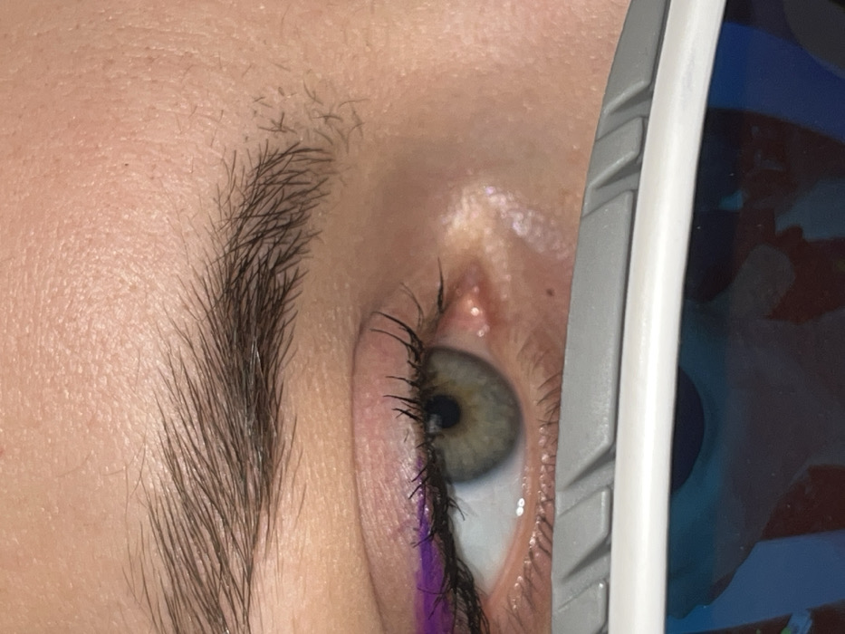 A close-up, high-resolution forensic image details the right ocular region of an individual in Krasnoyarsk, Russia. The skin displays a light to medium complexion with visible pores and minor textural variations across the forehead and periorbital area. A dark brown eyebrow with individual hairs is present above the eye, exhibiting a natural arch. The upper eyelid features a precisely applied line of vivid purple eyeliner. The eyelashes are dark, appear to be coated with mascara, and curve upwards. The iris is a distinct hazel-green color, with a clear, centrally positioned dark pupil. The sclera is white, with a minimal visible reddish capillary near the inner canthus. The lower portion of the image reveals the upper edge of eyewear, characterized by a textured, matte grey top band over a smooth white frame. Below the frame, a highly reflective blue-tinted lens is visible, reflecting a distorted light-colored, possibly angular, shape against a darker blue background.