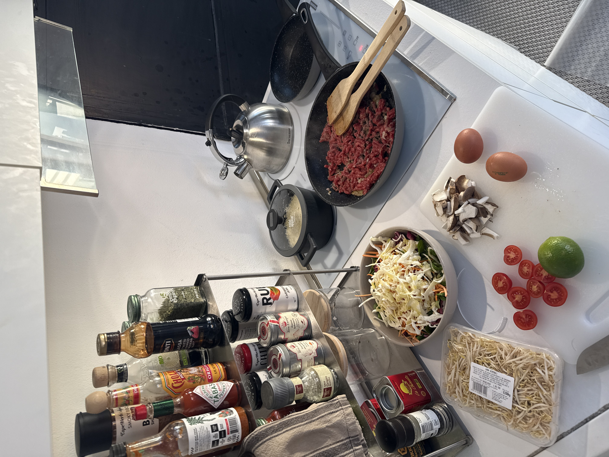 Here is a forensic description of the image:
**Scene Overview:** The image depicts a kitchen scene, likely in a residential setting, featuring food preparation in progress.
**Objects:**
* **Cooking Implements:**
* Multiple pans (one with ground meat being cooked)
* A kettle
* A pot with a lid
* Wooden cooking utensils (spatulas)
* **Ingredients:**
* Two raw eggs
* Sliced mushrooms
* A bowl of salad (cabbage, carrots, and other leafy greens)
* Sliced tomatoes
* A lime
* A package of bean sprouts
* **Condiments and Spices:** A rack filled with various bottles and containers of hot sauces, spices, and cooking oils (e.g., Tabasco, chili flakes).
* **Other:** A white cutting board, a gray kitchen towel.
**Environment:** The kitchen appears to be modern with white countertops and an electric stovetop. There is a black backsplash behind the stovetop. The lighting is bright.
**Location:** While the image does not provide specific information that would identify the location in Milan, Italy.