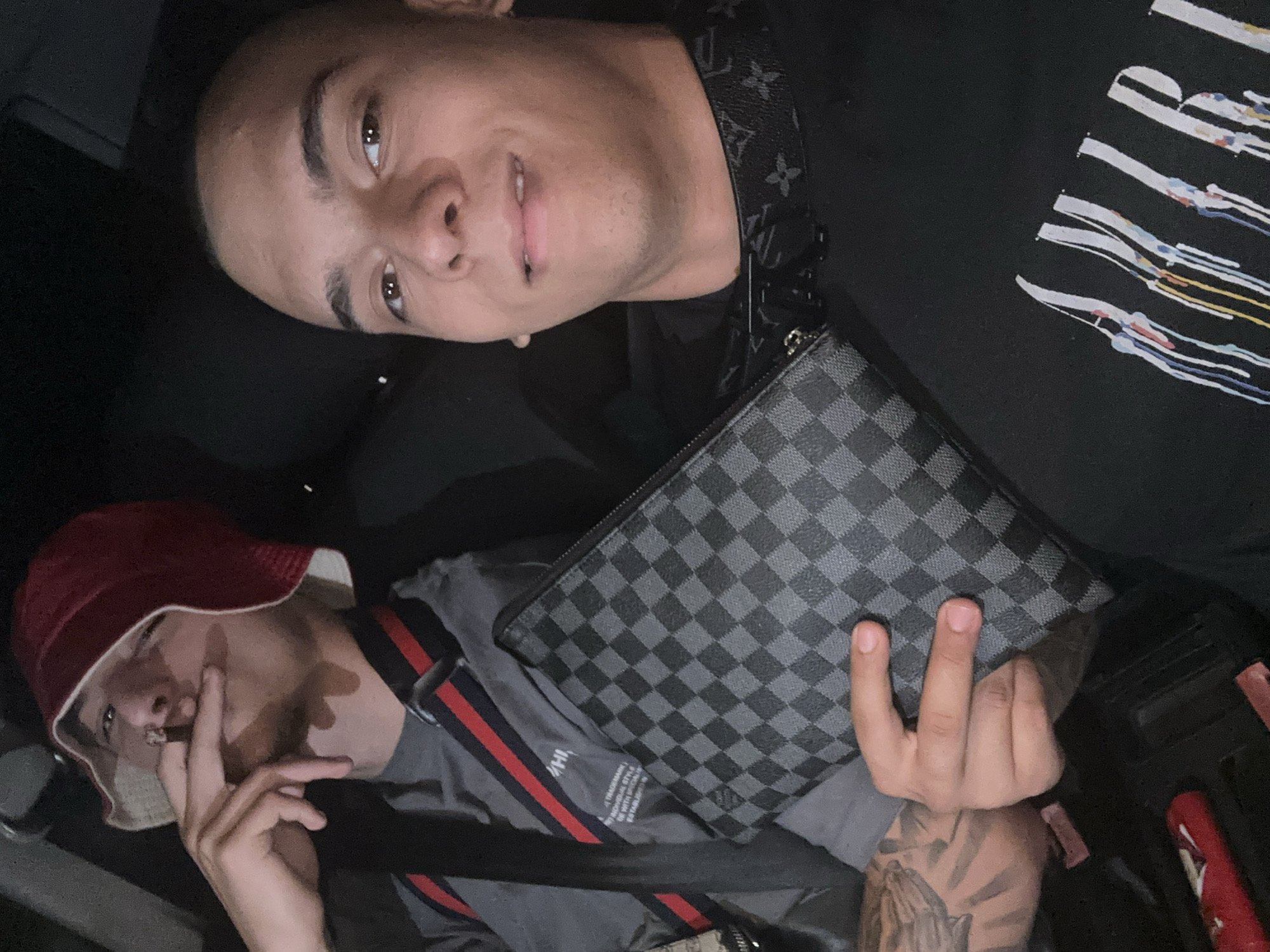 Two young men are in what appears to be a car. One man, positioned in the upper half of the image, has short, dark hair and is wearing a black shirt with white graphics. A Louis Vuitton patterned bag strap is visible across his chest, with a matching checkered pouch resting on his abdomen. The other man, in the lower half of the image, wears a red and white bucket hat and a gray shirt with red and navy stripes. He is smoking what appears to be a cigarillo. A Louis Vuitton checkered clutch or wallet lies nearby. The lighting is dim, suggesting it might be nighttime or the interior of a vehicle. There is no information to suggest the location being Plovdiv, Bulgaria.