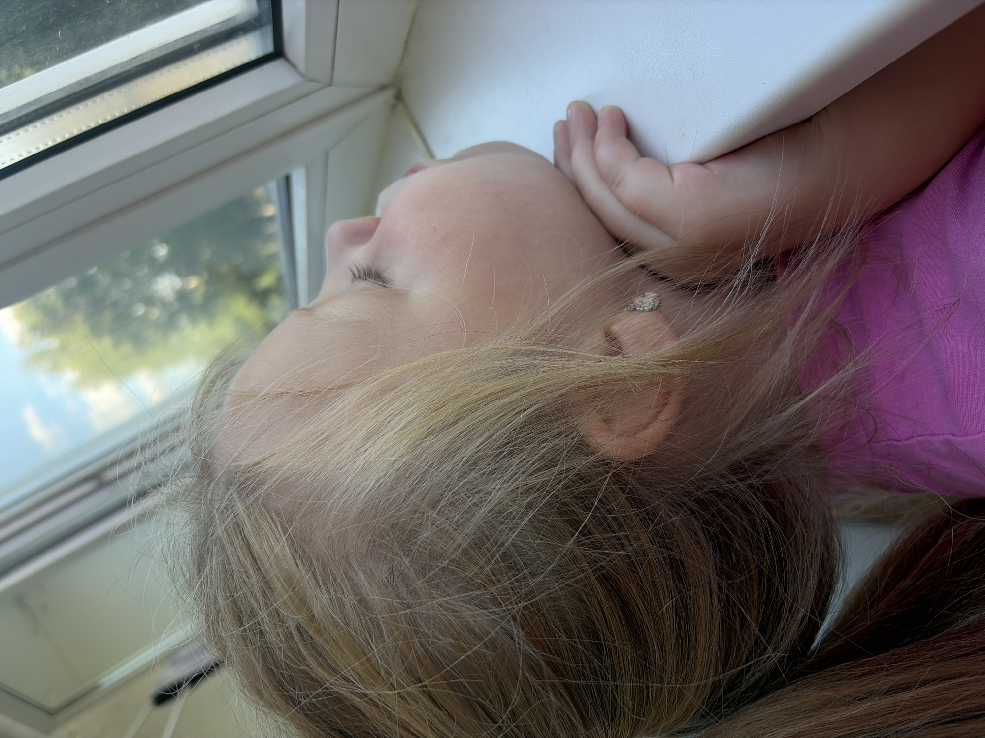 A young Caucasian girl with blonde hair is leaning on a white windowsill and looking out a window. She is wearing a pink shirt. Her left hand is supporting her chin. The girl has light skin and her eyes are closed. Outside the window, trees and a blue sky are visible. The scene appears to be indoors, likely in a residential setting. There is no information in the image that specifies the location in Izhevsk, Russia.