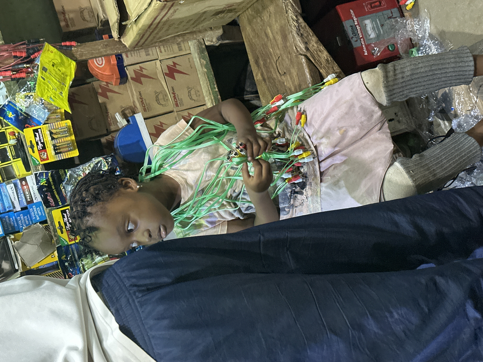 In Barden Yana, Nigeria, a young girl with braided hair sits amidst a cluttered market stall, her gaze directed to the left of the frame. She wears a light-colored t-shirt and a pink skirt, with grey leg warmers covering her lower legs. Her hands are occupied with a tangle of green wires, each tipped with colorful RCA connectors in red, yellow, and white. Behind her, shelves are stacked with various items, including boxes with "SOMelec" branding and a prominent red "Automatic Voltage Regulator" by Qlink. To the left of the frame, the dark, textured fabric of another person's clothing dominates the foreground, obscuring their identity. The overall impression is one of a busy, informal retail environment.