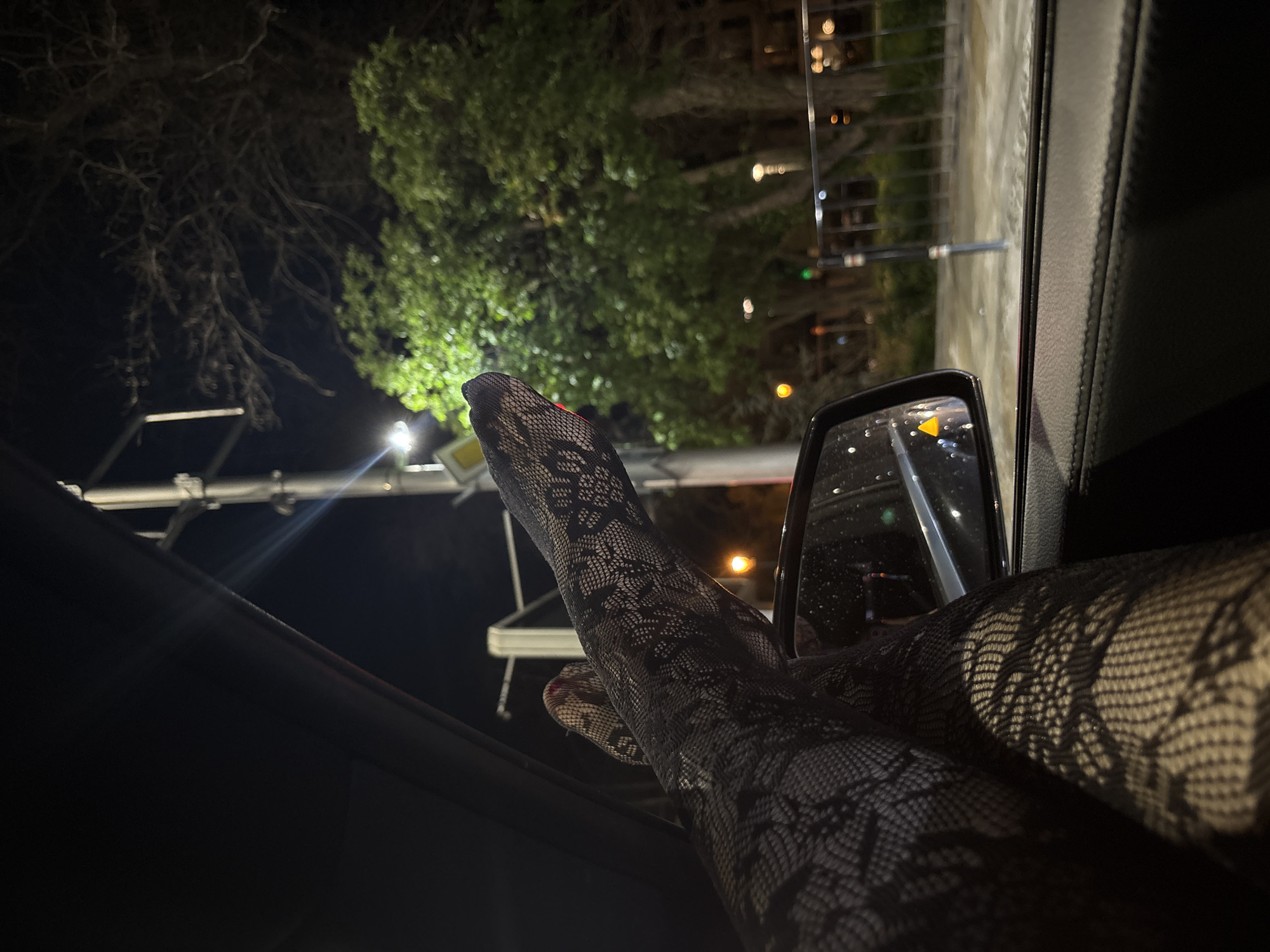 The photograph depicts two legs wearing black patterned hosiery extending from the interior of a vehicle towards the open window at night. The vehicle's side mirror is partially visible. Outside, the environment suggests an urban setting, with foliage from trees illuminated by streetlights. There are lampposts and what appears to be urban infrastructure visible in the background. The location is Plovdiv, Bulgaria.