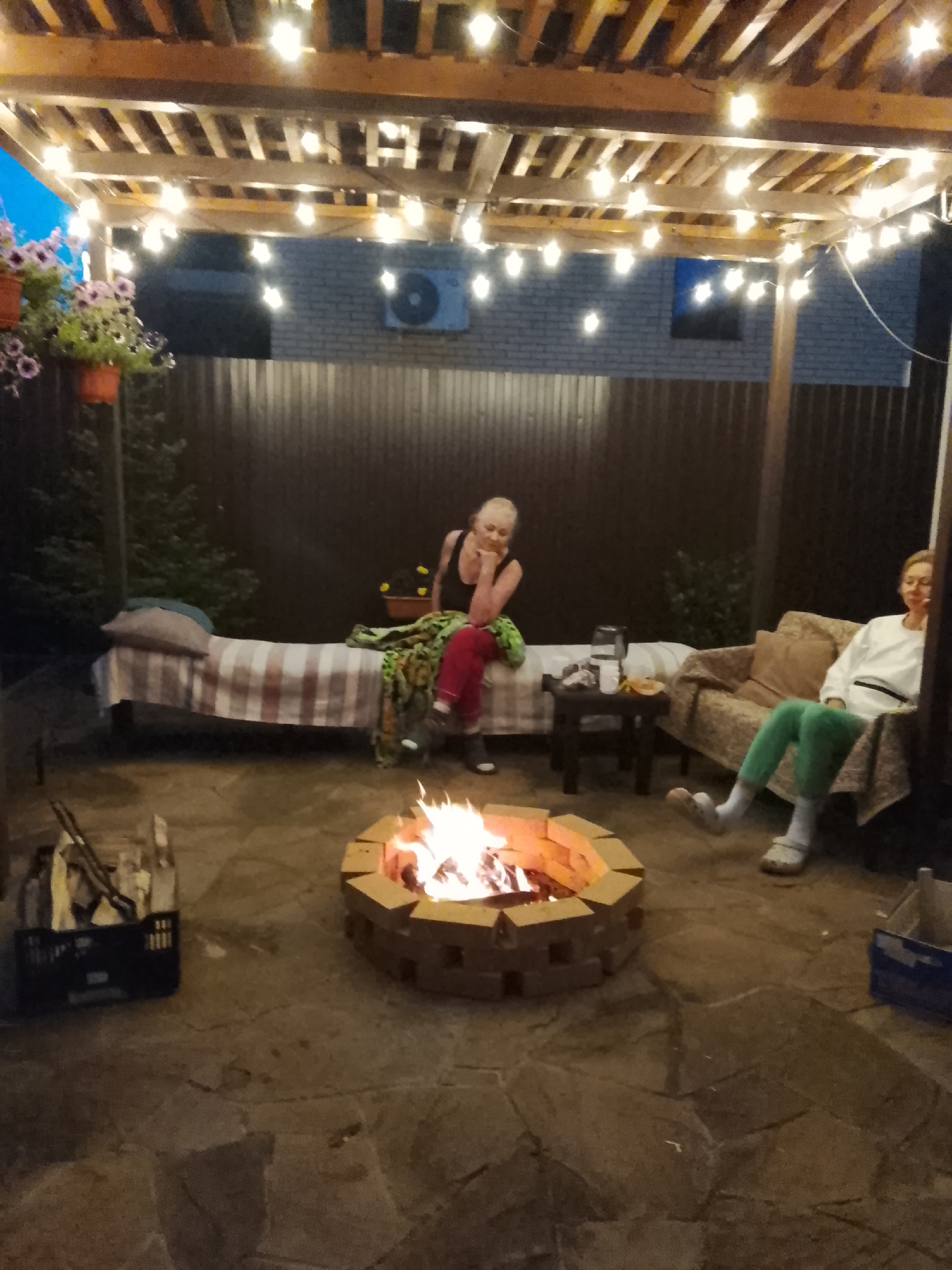 This is a medium shot of a backyard patio in the evening, likely in the village of Miniuno, Russia, illuminated by string lights under a wooden pergola and the glow of a brick fire pit. Two women are present. The woman on the left is sitting on a bed-like structure covered with a plaid blanket, leaning forward with her arms resting on her knees. She is wearing a black top and red pants. The woman on the right is seated on a patterned sofa, wearing a white long-sleeved shirt and green pants. She is leaning back and appears relaxed. In the foreground, the fire pit is actively burning. To the left of the fire pit, a black plastic crate contains firewood. The ground is paved with irregular stone slabs. In the background, a dark metal fence encloses the yard, and a brick wall with an air conditioning unit is visible. Hanging plants with pink flowers are on the left side of the image.