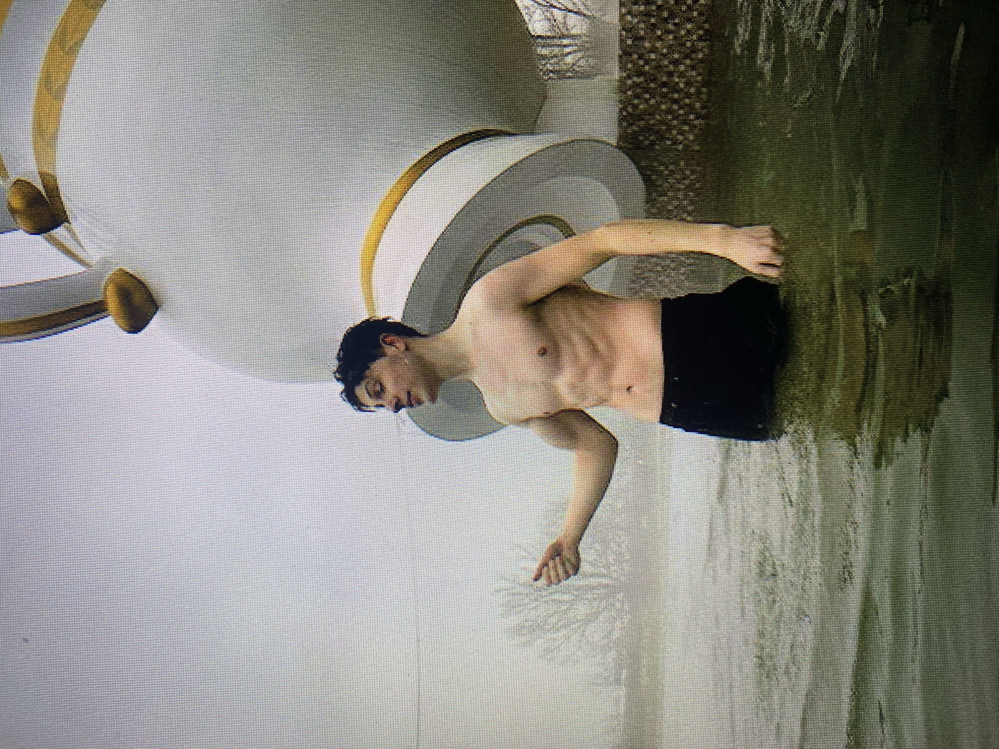 A young, shirtless man with dark hair stands in murky green water, wearing black shorts. His abdomen is muscular and defined. His left arm is extended forward, hand open. He is positioned in front of a large, white, bulbous structure with gold trim, resembling a teacup. The background is a hazy sky and bare trees, suggesting a cold or overcast day. The water appears to be a natural body, possibly a lake or river, as evidenced by the visible ripples and reflections. The overall impression is of a man participating in an outdoor activity, possibly a polar plunge, given the water's appearance and the man's attire. The structure in the background is a prominent landmark, likely artificial and decorative. This scene is located at Malta Lake in Poznan, Poland, specifically near the iconic "teacup" attraction.