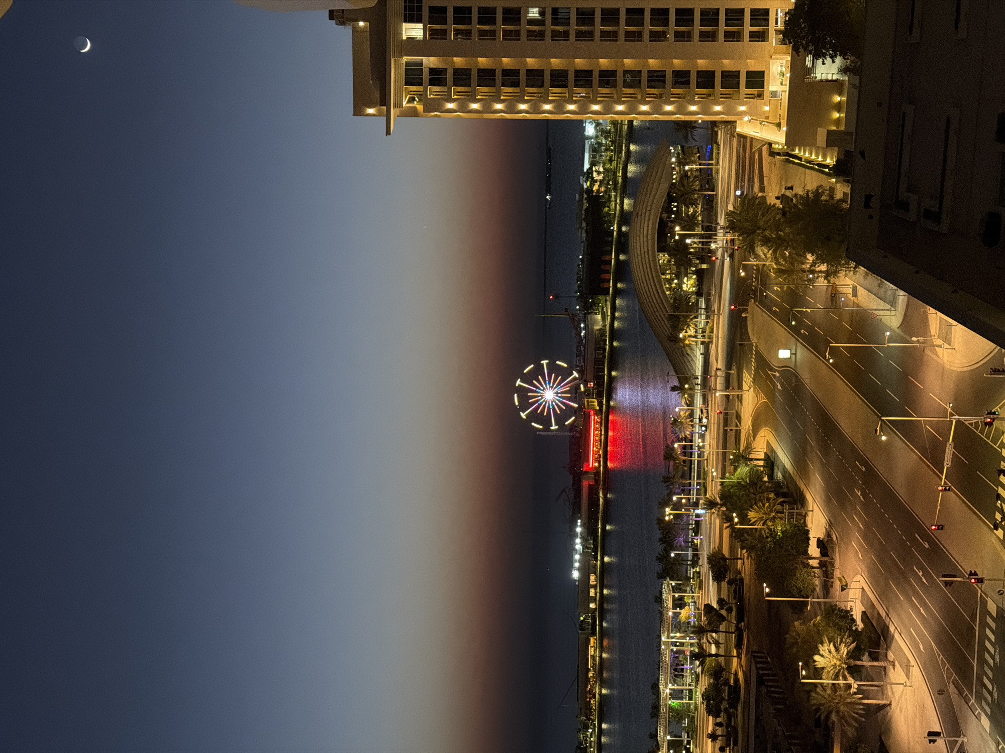 A nighttime aerial shot captures a city skyline with a body of water and a Ferris wheel. In the foreground, a multi-lane road is visible with streetlights and palm trees lining the sides. A tall, modern building with illuminated windows stands on the right side of the frame. In the distance, across the water, there are several buildings and structures, with the Ferris wheel, illuminated with colorful lights, prominently displayed. The sky above is a deep blue, transitioning to a gradient of orange and red near the horizon, indicating either dusk or dawn. A sliver of the crescent moon is visible in the upper right corner of the sky. The overall scene depicts a well-lit urban environment at night.
